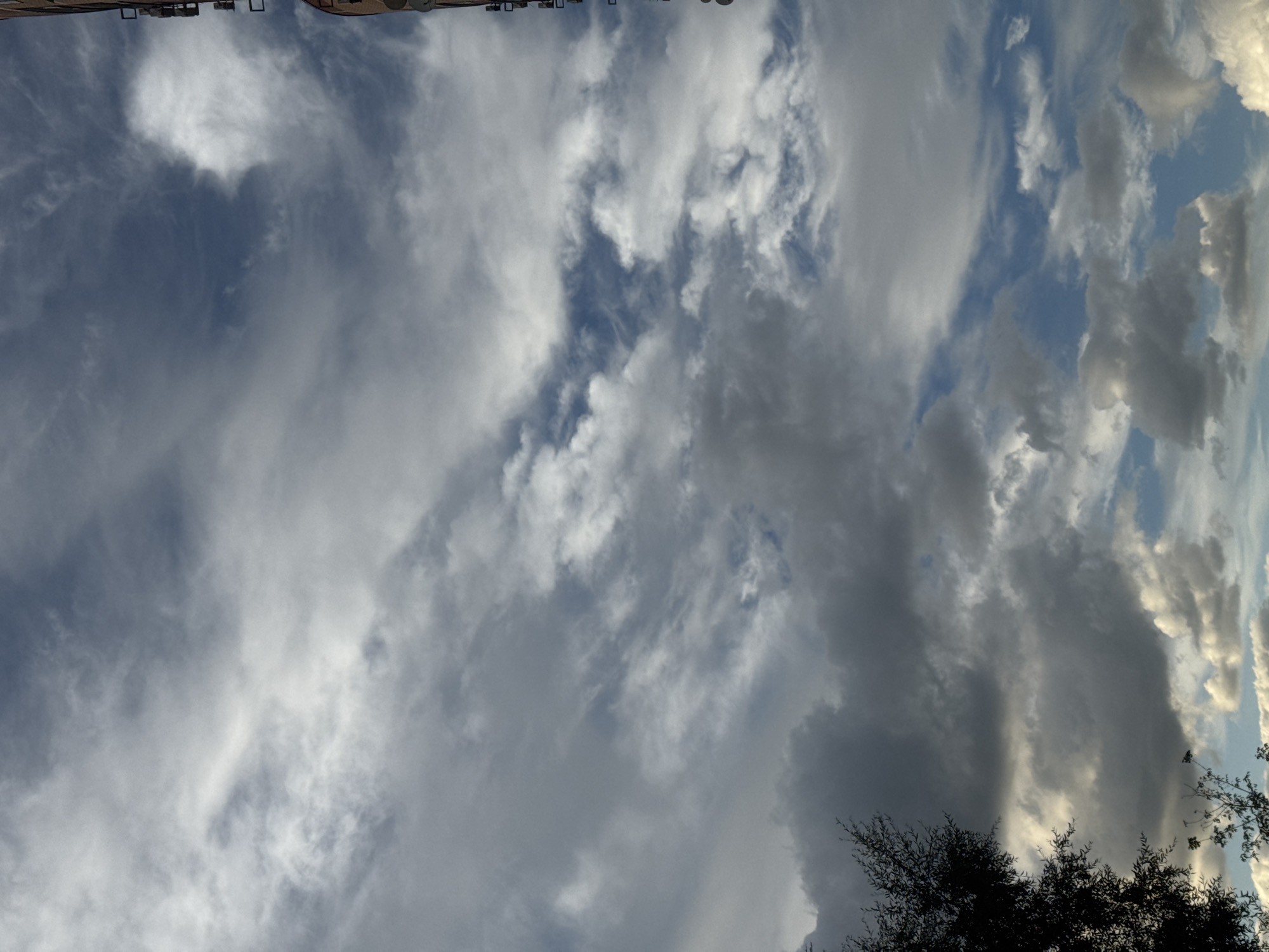 The image is a low-angle, full-frame shot of a cloudy sky, with the upper portion of a building visible on the right edge. The sky is filled with a mix of white and gray clouds, with patches of blue visible between them. The clouds appear to be moving, with some wispy formations and larger, more defined masses. On the right side of the image, the side of a multi-story building is visible, with what appear to be satellite dishes or antennas attached to its exterior. The building's facade is a muted, reddish-brown color. A silhouetted tree is visible on the left side of the frame, with its branches reaching into the cloudy sky. The overall impression is one of an overcast or partly cloudy day in an urban environment. The image does not contain any people, actions, or interactions. The location of Sovkhoza imeni Lenina, Russia is not discernible from the visual elements present in the image.