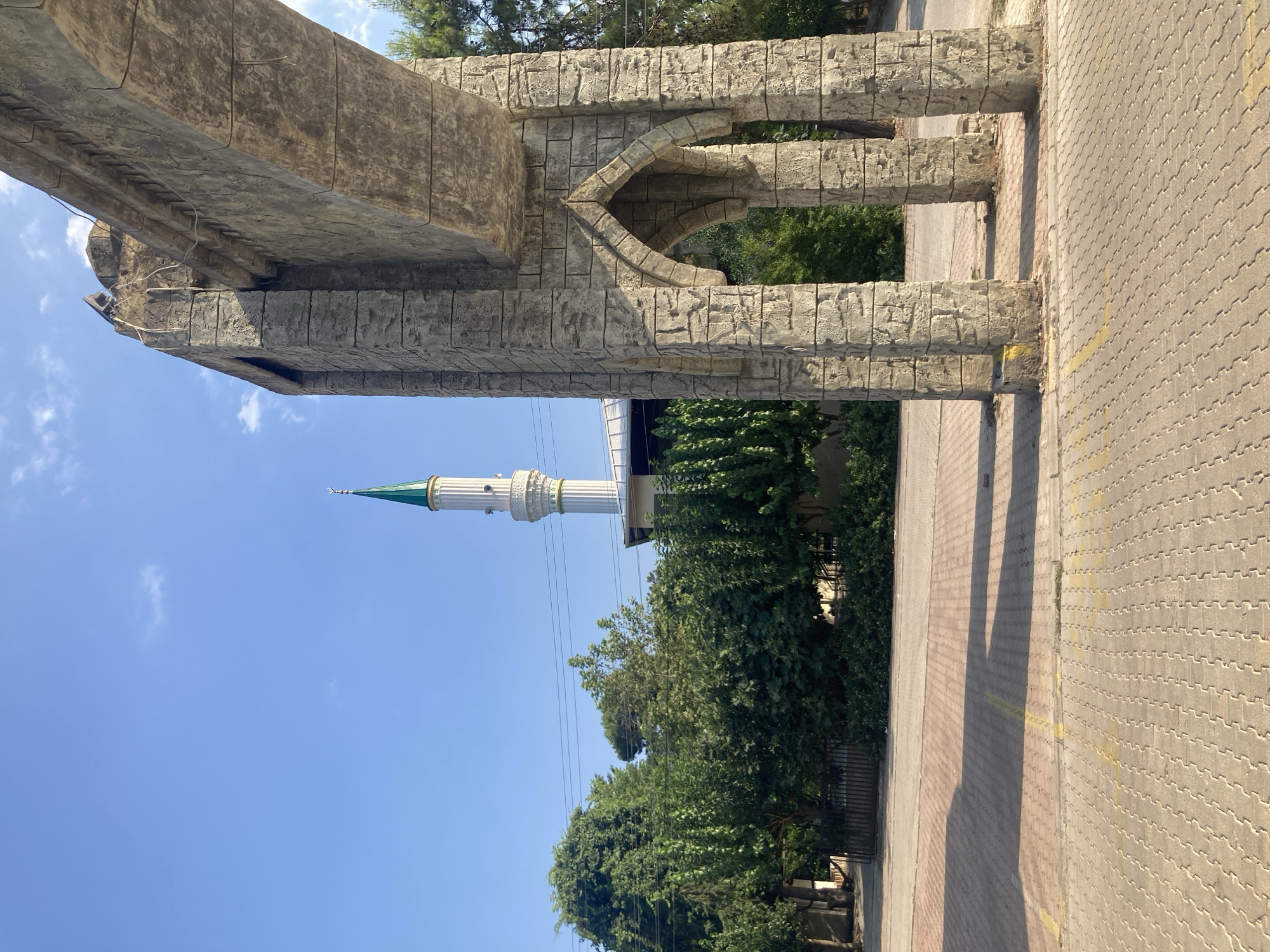 The image depicts an outdoor scene in Kemer, Turkey, dominated by a large stone archway in the foreground. The archway is constructed from rough, large stone blocks and features two arched openings. Behind the archway, a white minaret with a green spire rises against a clear blue sky, indicating the presence of a mosque. Lush green trees form a dense barrier between the archway and the mosque. The ground in the foreground is paved with interlocking concrete bricks, and a section of asphalt road is visible beyond the paved area. Sunlight casts long shadows across the paved ground, suggesting it is daytime. There are no people visible in the image.