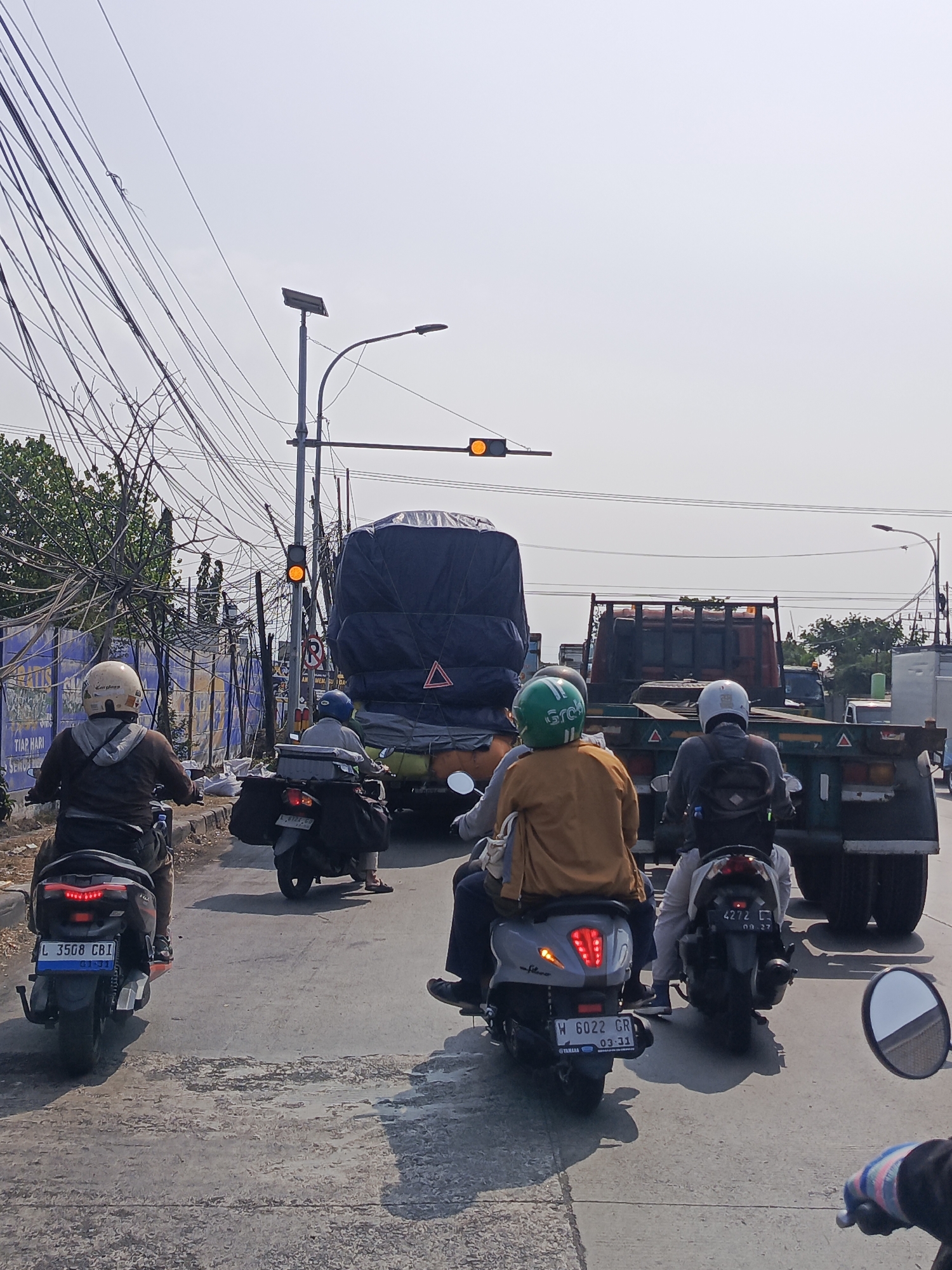 The image captures a traffic scene on a sunny day in Surabaya, Indonesia. Multiple motorcycles are present, with riders wearing helmets. A large truck, covered with a dark tarp, is visible in the middle of the road. Traffic lights are illuminated with yellow signals, suggesting a pause or transition in traffic flow. The environment includes power lines crisscrossing overhead and buildings lining the side of the road. The road surface is asphalt with visible markings and some wear and tear. The license plate on one of the motorcycles reads "L 3508 CBI".