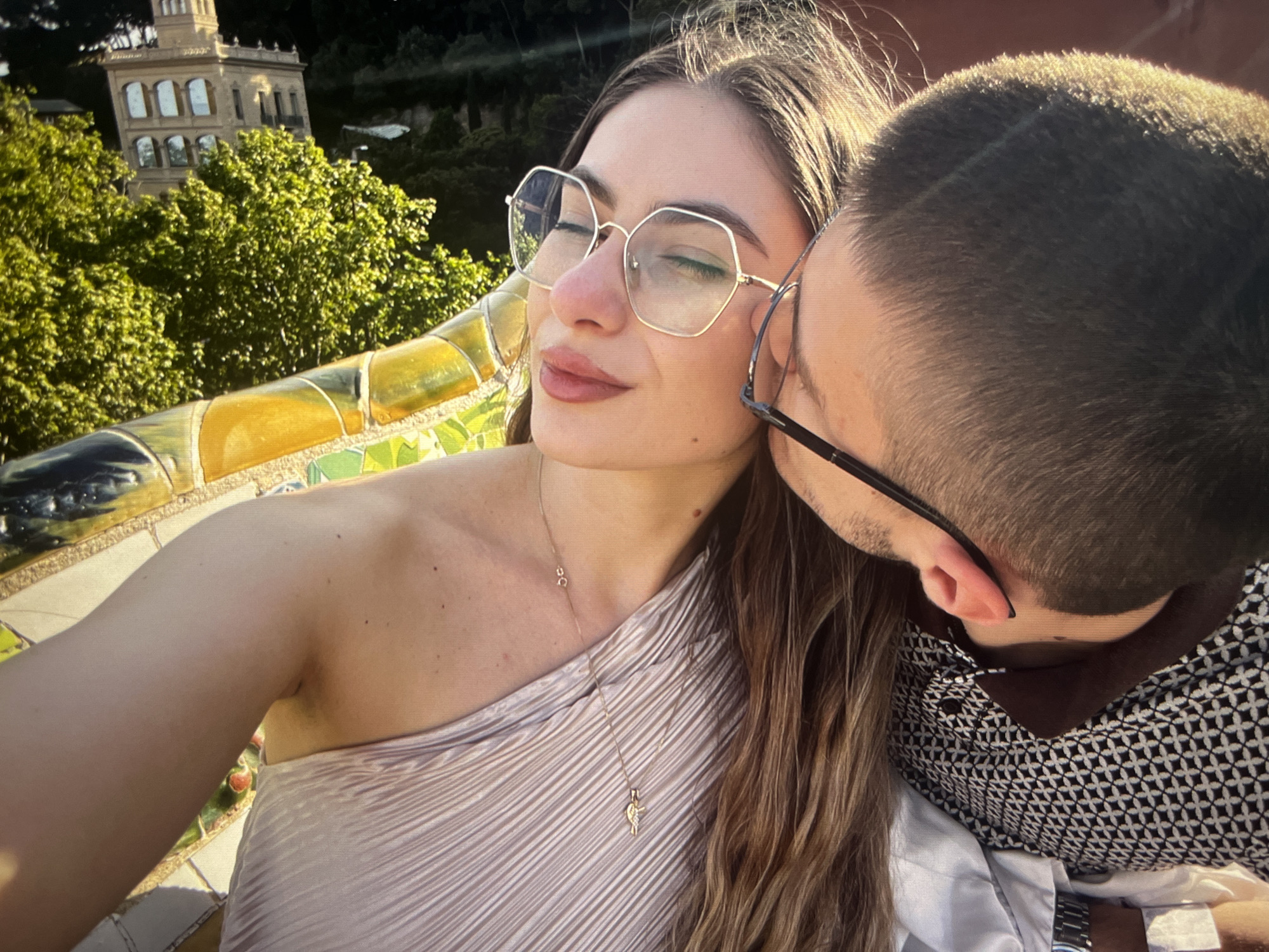 A young adult woman and man pose for a selfie outdoors. The woman is on the left, wearing a beige, one-shoulder dress and gold-rimmed, hexagonal glasses. Her eyes are closed, and she wears a gold necklace with a small pendant. The man, on the right, is wearing glasses and a patterned shirt. He appears to be kissing the woman’s cheek. The background includes a building with multiple arched windows, trees, and a mosaic-tiled bench or wall. The location appears to be Park Güell in Barcelona.