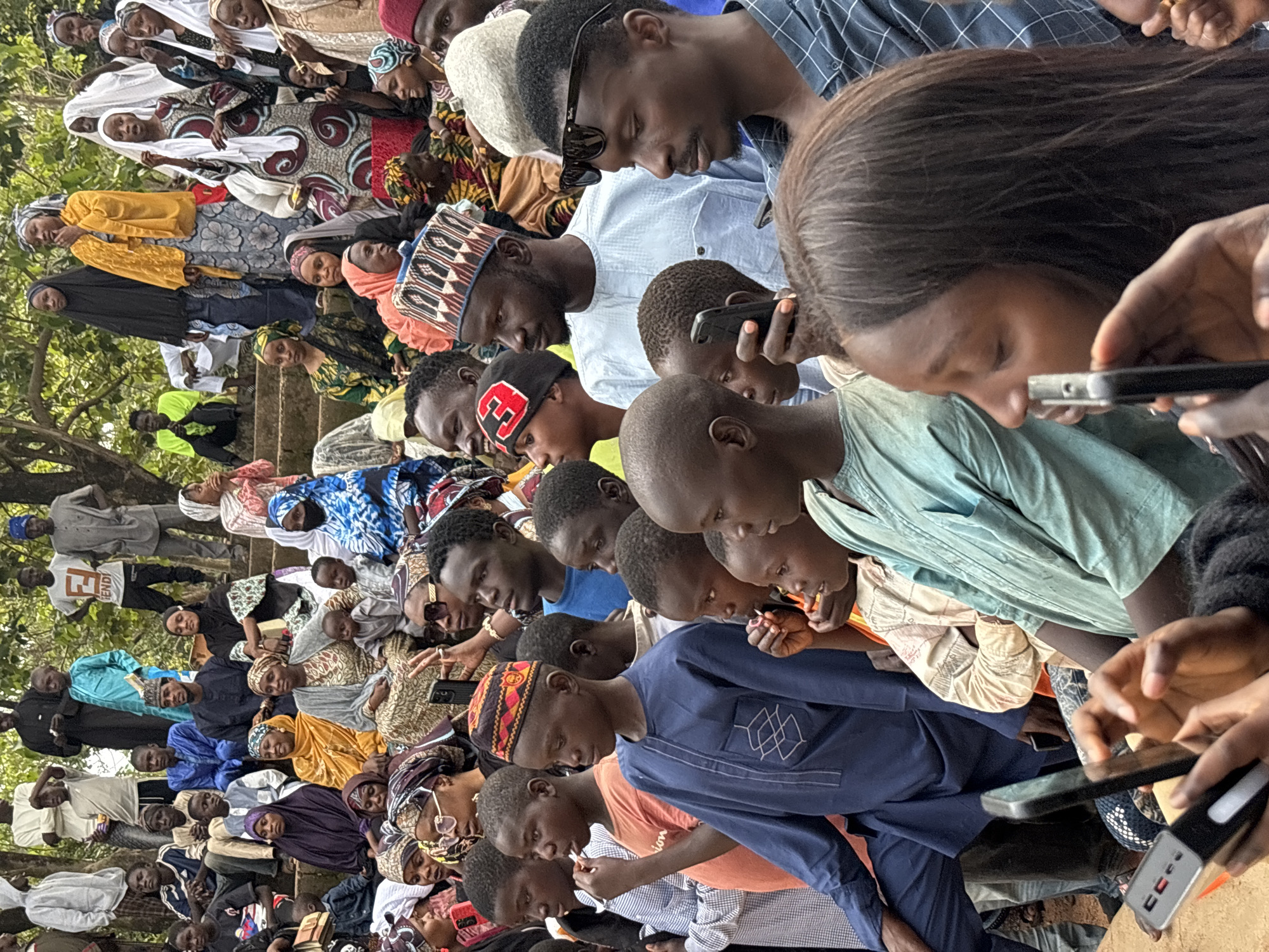 The image depicts a crowd of people, appearing to be in an outdoor, possibly dusty environment, with trees and foliage visible in the background. The people are dressed in a variety of traditional and modern clothing, including headscarves, turbans, and caps. Many are looking in the same direction, suggesting they are observing an event or performance. Several individuals are holding up their mobile phones, possibly to take pictures or record video. The atmosphere seems to be one of gathering and observation, with a mix of ages present. The scene could potentially be a festival, a cultural event, or a public gathering. It is not possible to determine the specific location in the city of Jos, Nigeria from the image alone.