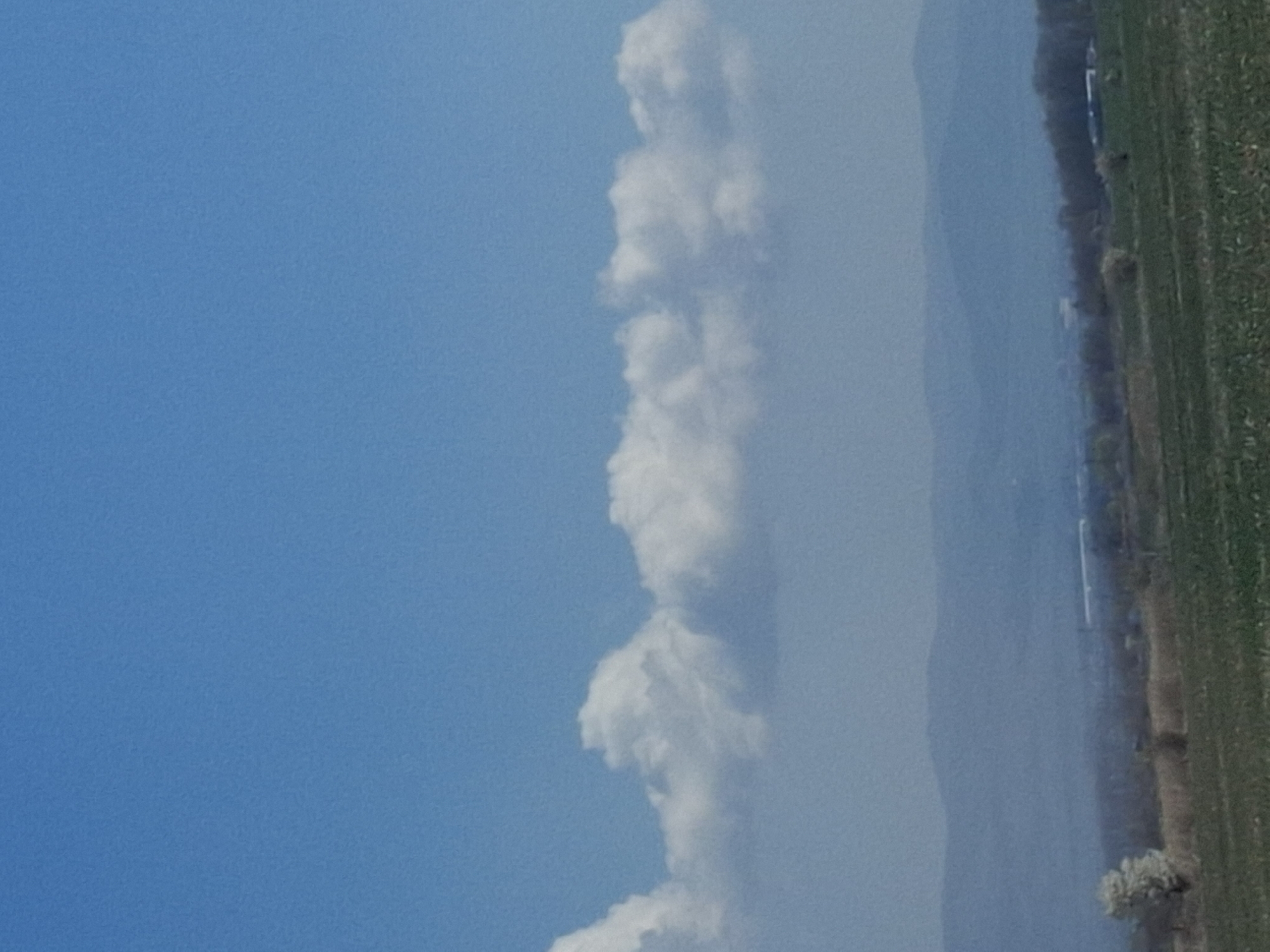 The image shows a view of the sky over Krivina, Bulgaria. A column of cumulus clouds rises vertically in the middle of the frame. The sky is blue, and on the right of the frame, there is a field of green grass with a line of trees and vegetation along a body of water. The water appears to have mist above it.