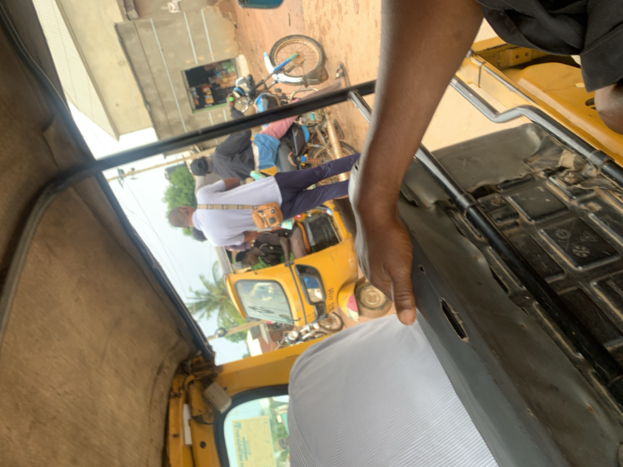 The image is taken from inside a yellow auto-rickshaw, looking forward and to the right. The interior of the auto-rickshaw has a worn, light-colored fabric ceiling and a gray upholstered seat in the foreground. A person's dark-skinned hand with short fingernails is resting on the edge of the seat. Outside, the auto-rickshaw is stopped on a dirt road in what appears to be a commercial area. Another yellow auto-rickshaw is parked next to it, with a person sitting in the driver's seat. Two men are standing behind the parked auto-rickshaw. One man is wearing a light blue polo shirt and a brown messenger bag slung over his shoulder. The other man is wearing a black jacket and a black cap. They are facing away from the camera. To the right of the parked auto-rickshaw, a motorcycle is visible, with a person wearing pink pants and a black jacket riding it. In the background, there are buildings, including a store with shelves of goods displayed in its open front. Palm trees are also visible in the distance. The scene suggests a busy street in Karu, Nigeria, possibly during the day.