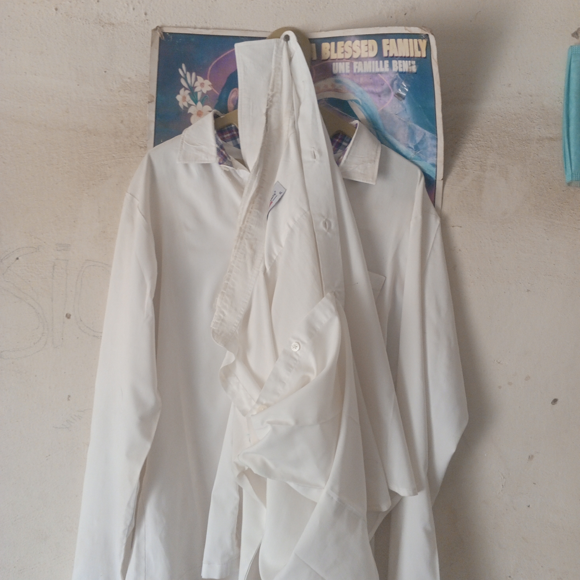 A white collared shirt and a blue face mask are hanging on a textured, off-white wall. The shirt is draped over a hanger and appears slightly rumpled, with a plaid pattern visible on the inside collar. A poster featuring a floral design and text that reads "BLESSED FAMILY" and "UNE FAMILLE BEN" is tacked to the wall behind the shirt. Faint, sketched markings are visible on the wall to the left of the shirt. The scene is likely an outdoor setting, possibly in Zinna, Nigeria, given the texture of the wall and the presence of the poster.