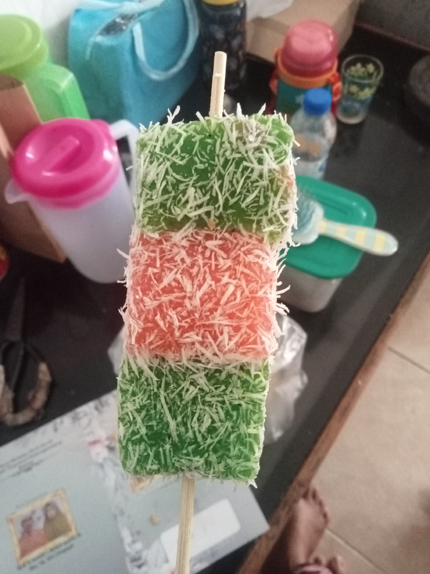 The image shows a close-up of a layered dessert on a stick, likely a type of traditional Indonesian sweet. The dessert has three distinct layers: green on top, red in the middle, and green on the bottom. Each layer is coated with shredded white coconut. The dessert is positioned vertically on a dark, reflective surface, which appears to be a countertop.
In the background, several objects are visible, suggesting a domestic or casual setting. To the left, there are several plastic containers with colorful lids, including a pink-lidded one and a green one. Further back, a bright blue container with a handle is partially visible. To the right, a small glass with a floral pattern and a clear plastic bottle with a blue cap are present. A green food container with a spoon or utensil inside is also visible on the right side of the image. In the lower left, a paper with a framed picture of two people is partially visible. The flooring in the lower right is tiled. A person's bare foot is also visible in the lower right corner of the image.
While the image itself does not provide specific landmarks to pinpoint the exact location within Malang, Indonesia, the presence of traditional Indonesian desserts and common household items suggests a scene typical of Indonesian domestic life. Malang is a city in East Java, Indonesia, known for its pleasant climate and cultural heritage. The objects and food in the image are consistent with what might be found in a home or a local market in Malang.