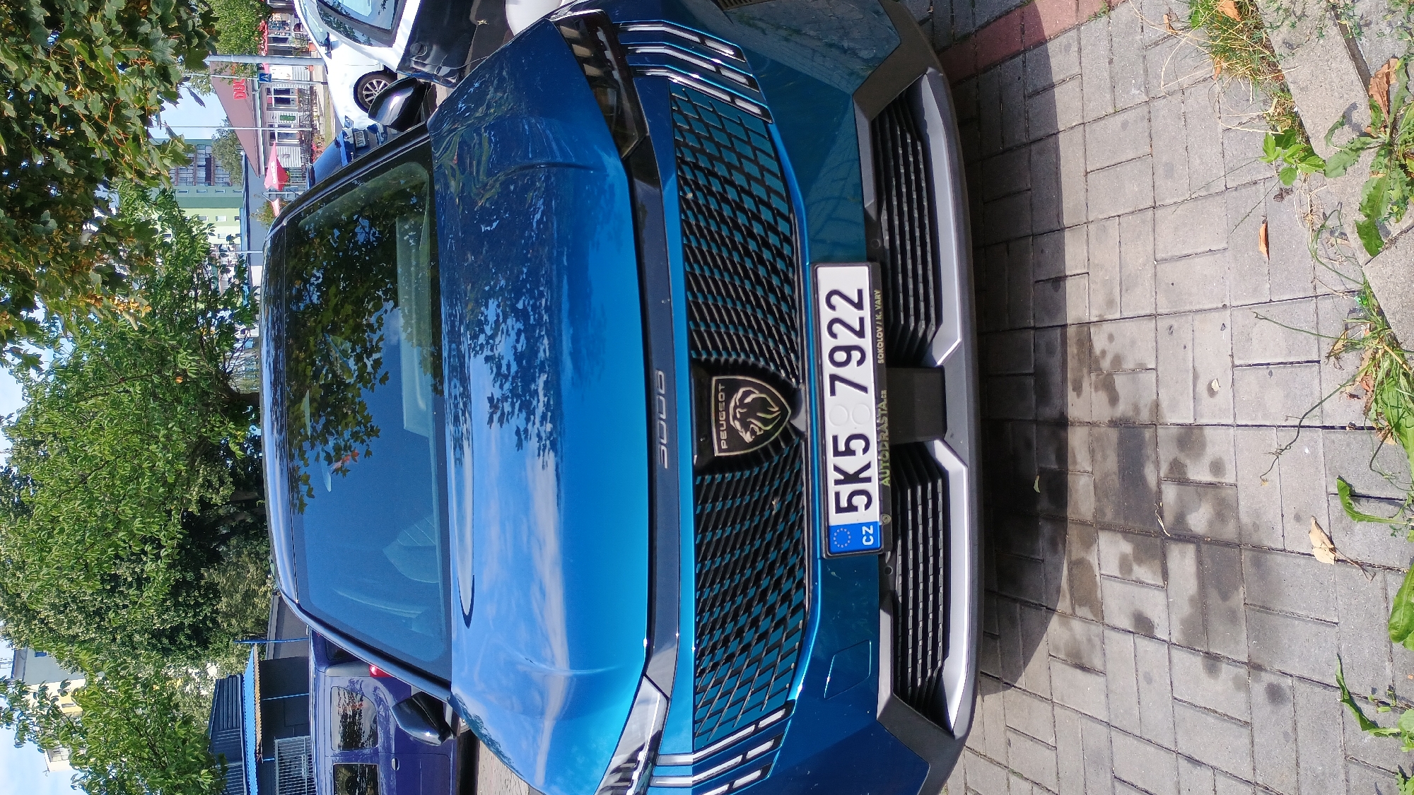 An eye-level, medium shot captures the front of a blue Peugeot 3008 parked on a grey cobblestone surface in Sokolov, Czechia. The car's grille is black with a diamond pattern and a silver Peugeot logo in the center. The license plate reads "5K5 7922" with "CZ" on the left and "AUTODRASTA.CZ SOKOLOV / K. VARY" on the bottom. In the background, trees with green foliage obscure parts of apartment buildings and a red building with a white sign. The sky is visible through the leaves and clouds are reflected in the car's windshield. The ground shows water stains and scattered leaves.