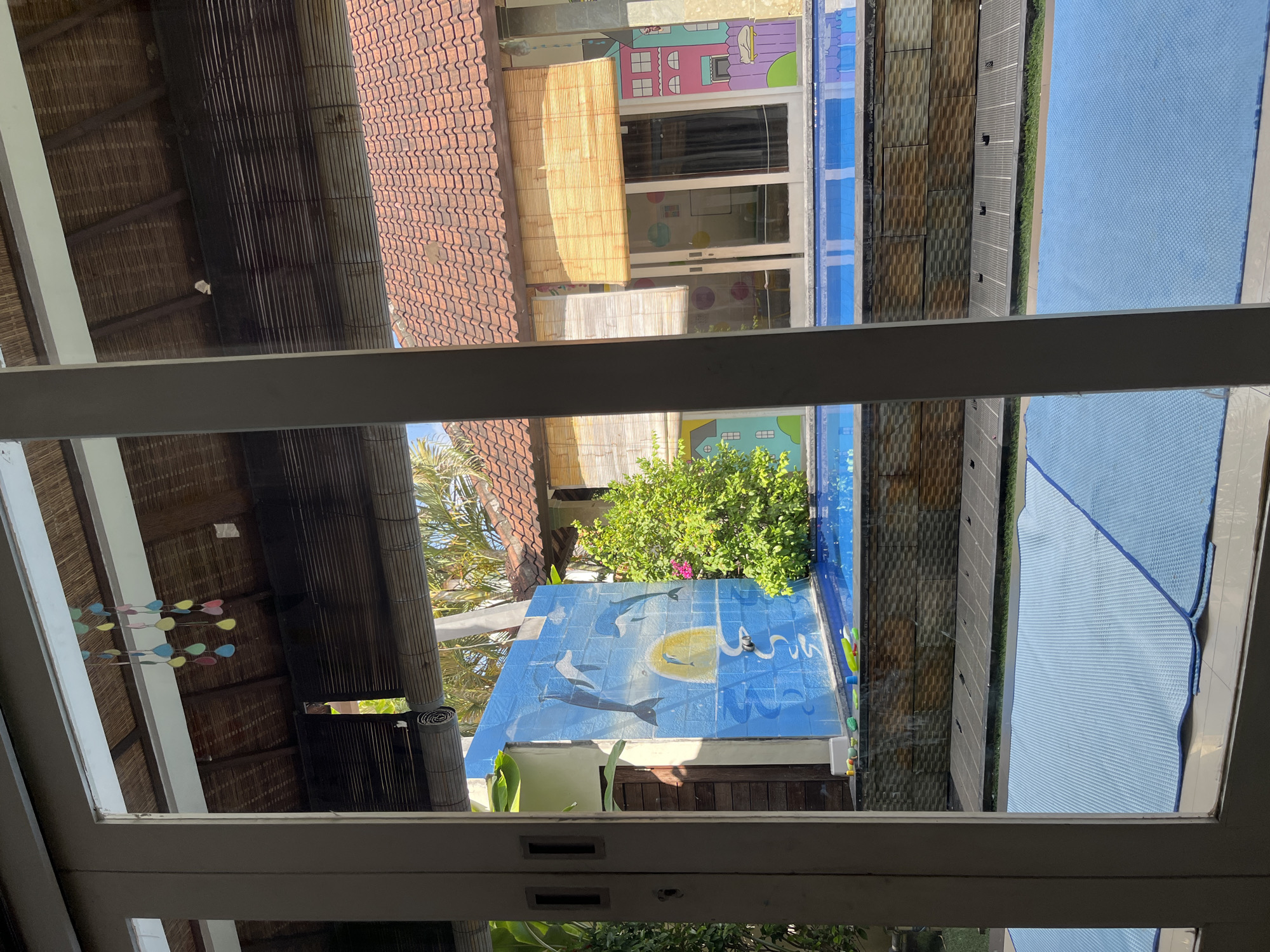 This image depicts an outdoor swimming pool area, likely in Kuta, Indonesia, as suggested by the architectural style and tropical foliage. The scene is viewed through a window frame with white pillars on either side.
In the background, a bright blue swimming pool is visible, surrounded by a textured wall made of stone-like tiles in earthy tones of brown and gray. To the left of the pool is a vibrant mural painted on a light blue wall, featuring stylized dolphins jumping out of the water, with a yellow sun setting behind them. A small, lush green tree with pink flowers stands beside the mural, its branches reaching towards the pool.
Further back, buildings with orange tile roofs are visible. One building has large windows covered by bamboo blinds, and another features colorful cartoon-style drawings of houses on its exterior walls. Palm trees and other tropical vegetation are interspersed throughout the background, indicating a warm climate.
In the foreground, a large area covered with blue mats, possibly for lounging or exercise, stretches across the bottom of the frame. There are also some children's toys scattered near the edge of the pool. The overall impression is that of a relaxed, resort-like environment. The lighting suggests it is daytime, with bright sunlight casting shadows. There are no people visible in the image.
