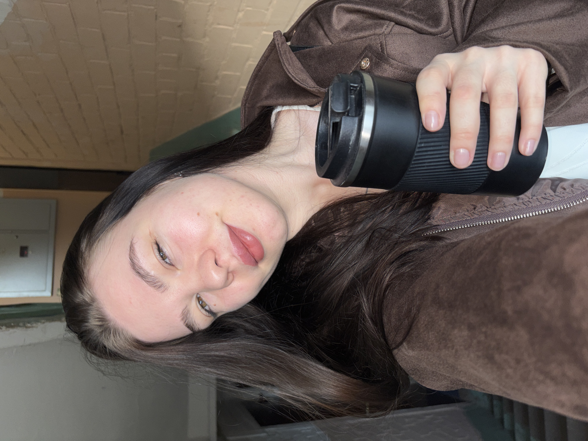 A woman is taking a selfie indoors. She has light skin, brown hair, and is wearing a brown jacket over a white top. She is holding a black travel mug in her right hand. The background includes a cream-colored brick ceiling and a gray wall with a utility box. The image does not provide enough information to determine the location in Perm, Russia.