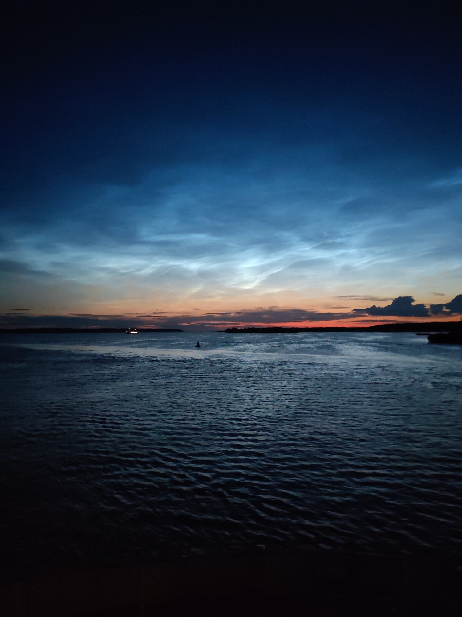 This is a long shot, horizontal, eye-level, outdoor, full-color, daytime photograph of the Rybinsk Reservoir in Rybinsk, Russia. The sky is a deep blue, transitioning to lighter blues and oranges near the horizon. Wispy clouds are visible in the sky, illuminated by the setting sun. The reservoir's surface is dark and choppy, reflecting the sky's colors. A faint silhouette of a boat is visible in the distance, as well as a buoy. The shoreline is a dark, indistinct line in the distance. The foreground is dark, with no visible subjects or objects. The overall mood of the image is serene and peaceful.