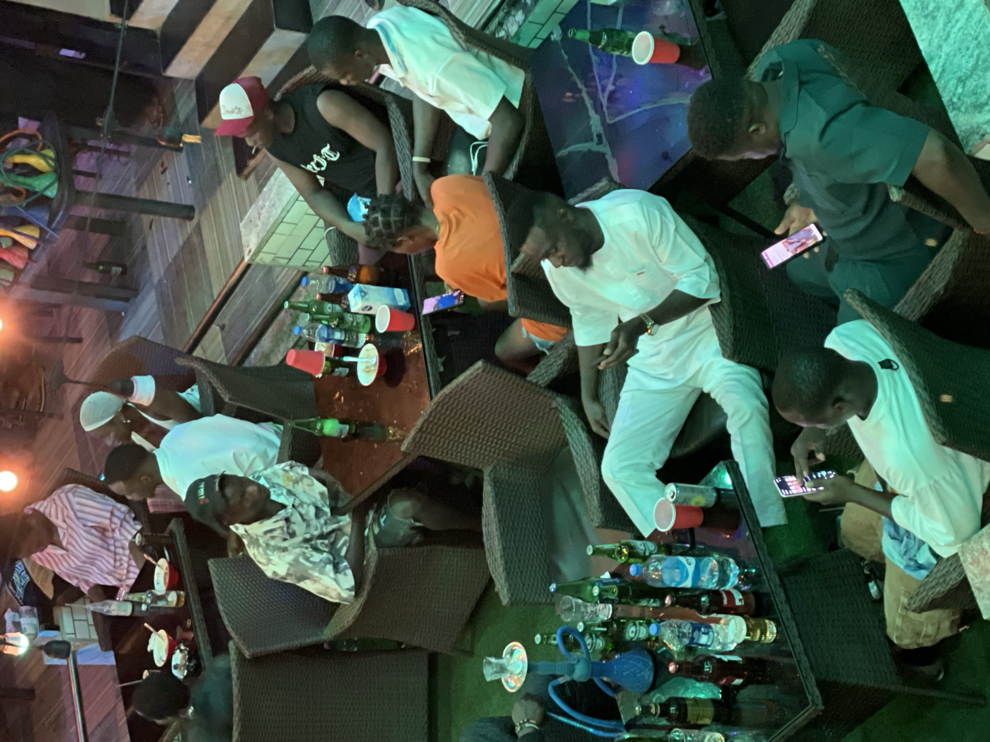 The image depicts a group of about eight men gathered at a table at a restaurant, possibly in Obosi, Nigeria, based on environmental context. The men are seated around a brown table with a reflective surface. They are casually dressed, wearing shirts of various colors, including white, orange, green, pink, and black, and some are wearing hats. Several are looking at their mobile phones. The table is crowded with plastic cups, glass bottles, and bottled water. A hookah is also on the table. The setting appears to be a patio or outdoor area with woven chairs. A staircase is visible in the background.