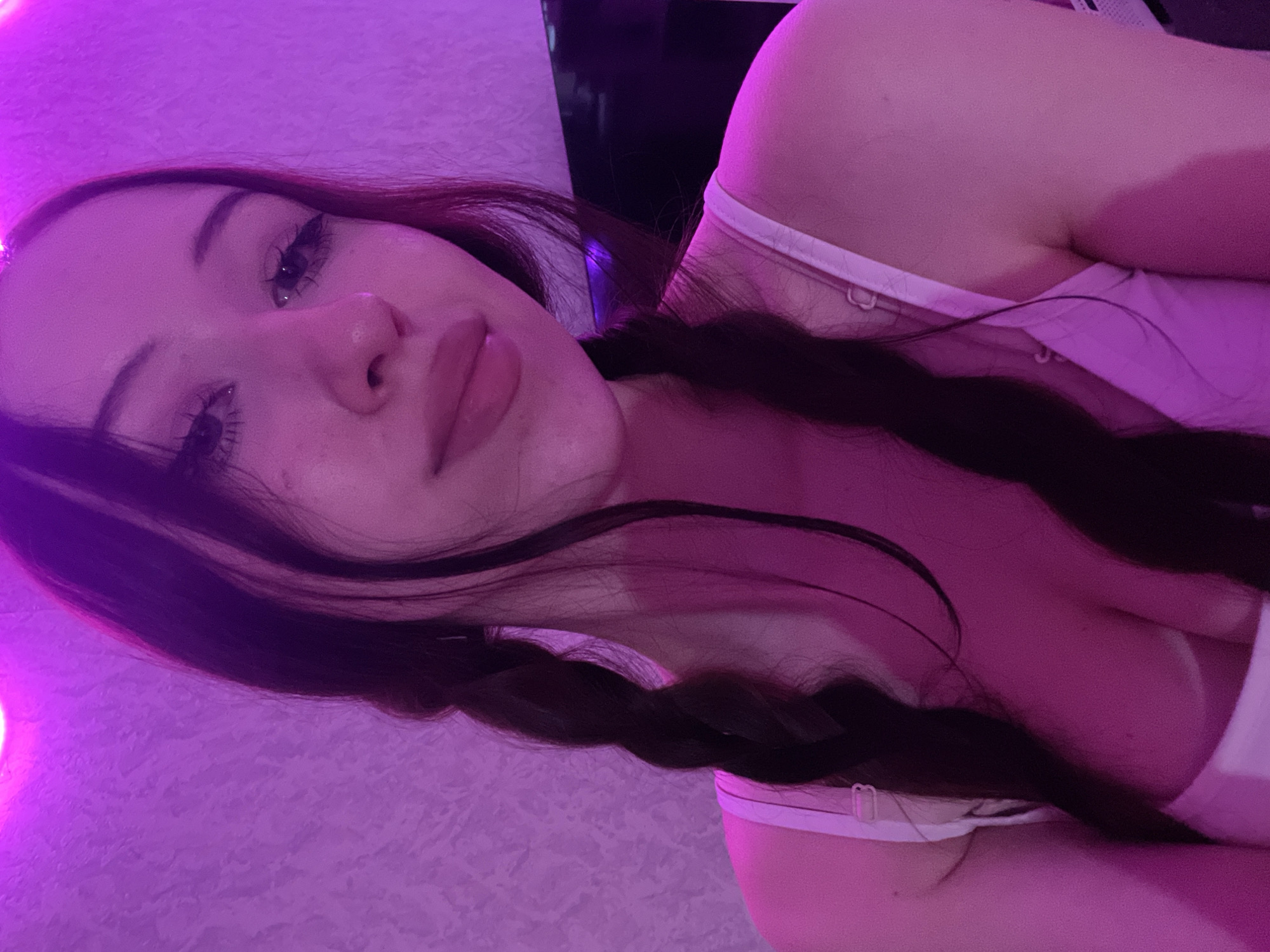 The image features a young woman positioned indoors, located in Votkinsk, Russia. She has long, dark hair styled in two braids, which rest over her shoulders and chest. Her gaze is directed towards the viewer, with distinct eyeliner visible. Her lips appear full. She is wearing a white or light-colored garment, possibly a bra or camisole, with straps visible on both shoulders and across her upper chest. The scene is illuminated by a dominant magenta-purple light source, casting this hue over her skin and the textured wall in the background. A dark, indistinct object, possibly furniture or electronics, is partially visible on the right side of the frame.