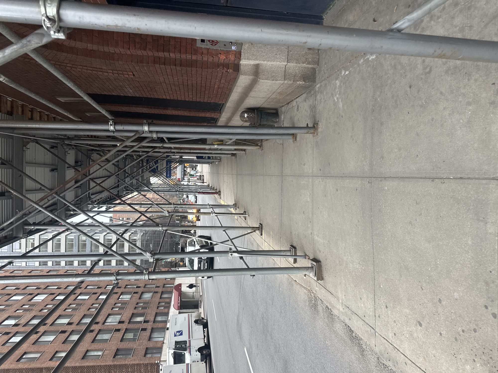 The image depicts a street scene on a sidewalk covered by extensive metal scaffolding in New York City, United States of America, specifically near 200 East 30th Street in the Murray Hill neighborhood.
The perspective is from the sidewalk, looking down the street. The foreground is dominated by grey metal scaffolding, consisting of numerous vertical poles, horizontal braces, and diagonal cross-braces, supporting an overhead structure. This scaffolding extends along the entire right side of the image, obscuring the upper portion of a brick building with a grey stone base and dark arched entrances. A dark-colored public trash can with white graffiti is visible beneath the scaffolding on the right, positioned against the brick wall. A silver utility panel is mounted on the brick wall further right.
To the left of the scaffolding, a multi-lane asphalt street runs parallel to the sidewalk. A white United States Postal Service (USPS) truck, marked with "United States Postal Service" and "We Deliver For You" logos, is parked on the street's left side, facing away from the camera. Behind the USPS truck, a pedestrian walks away from the camera under a red awning, which partially covers an entrance labeled "Jan Tus" or similar text. Directly across the street from the parked truck, a white SUV is in motion, driving towards the viewer. Further down the street, additional vehicles and indistinct pedestrians are visible.
The left side of the street is lined by multi-story buildings. A large, reddish-brown brick apartment building with numerous windows occupies the immediate left, while a taller, light-colored building with a grid of windows is visible in the distance. The sky appears overcast, providing diffused daylight illumination. A blue street sign, partially visible under scaffolding on the left, indicates "Murray Hill" and "200 East 30th Street." The concrete sidewalk shows signs of wear and minor staining.