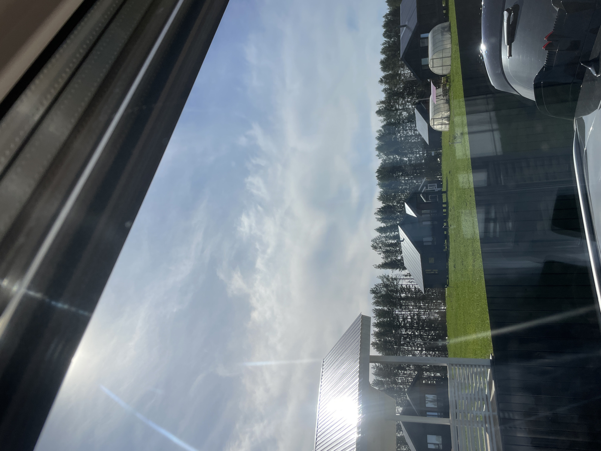 The image shows a view from a window. Outside, there are several dark-colored buildings situated among trees. There is a patch of green lawn. The sky is visible with some clouds. Part of a car is reflected in the window. The location is unknown.