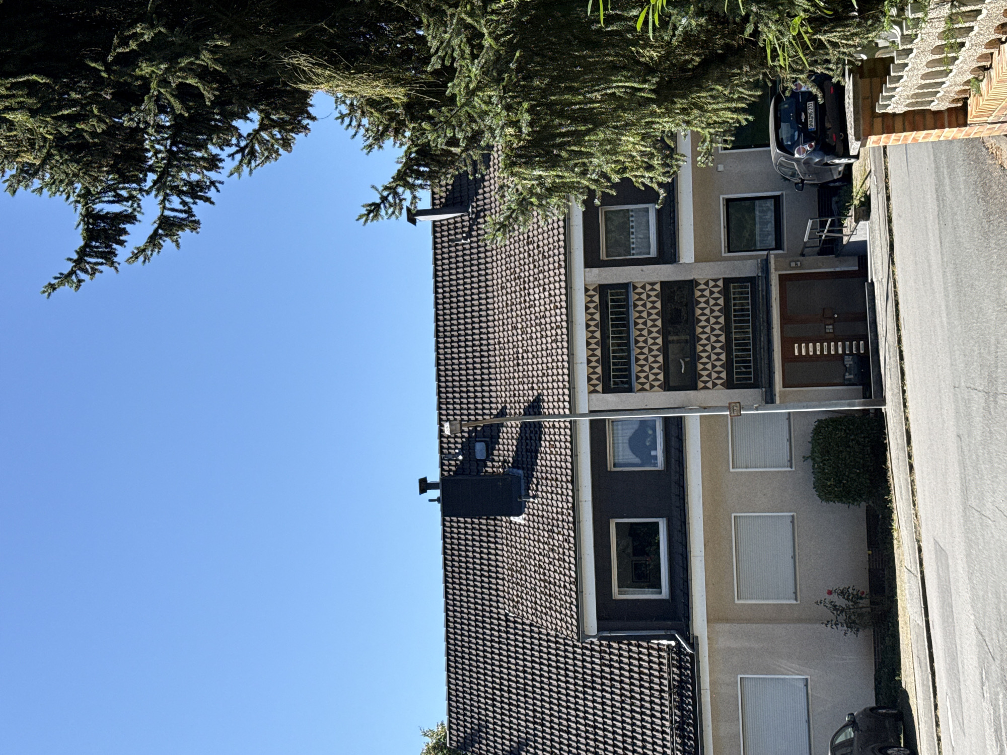 The image shows a multi-story building with a dark tiled roof, under a clear blue sky. The building features several windows, some with roller shutters and others with visible interiors. A dark chimney protrudes from the roof. The facade of the building is a mix of light beige and dark gray sections, with a patterned decorative element on one of the upper floors. A dark colored car is parked in front of the building. A large evergreen tree with dense foliage partially obscures the right side of the building and the sky. The building is situated on a street with a sidewalk and a paved road. In the foreground, a brick fence is partially visible. The scene is a street view in the city of Bad Sachsa, Germany.
