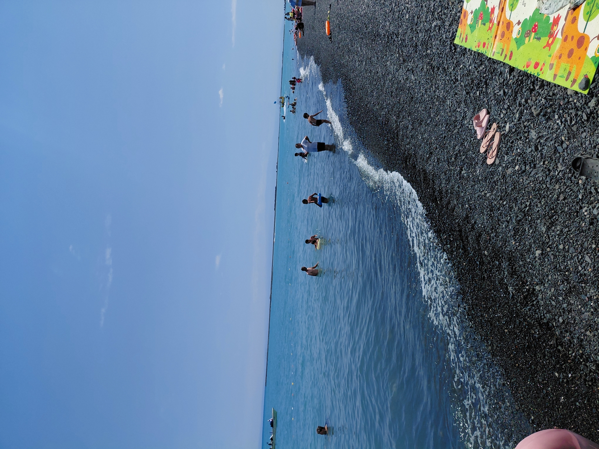 The image depicts a sunny day at a pebble beach on the shore of Lake Balkhash in Ақши, Kazakhstan. Several people are visible in the shallow, turquoise water, including children playing and adults wading. Some are swimming, and one person appears to be on a yellow float. A boat is visible in the distance on the lake. The beach is covered in dark gray pebbles, and a colorful mat and a pair of pink flip-flops are placed on the shore. The sky is clear and blue with a few wispy clouds.