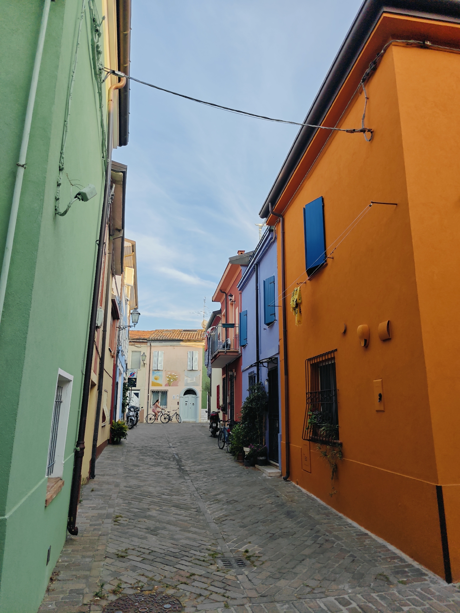 The image depicts a narrow, cobblestone street lined with brightly colored buildings in Rimini, Italy. The buildings on either side of the street are painted in vibrant hues of orange, green, and purple, creating a striking visual contrast. The architecture suggests a Mediterranean setting.
The scene appears to be captured in the late afternoon or early evening, with the warm sunlight casting shadows and highlighting the textures of the buildings and the cobblestones. The sky above is a clear blue with wispy white clouds, indicating pleasant weather.
In the distance, two people are visible on bicycles, navigating the street. One person, dressed in pink, is riding a bicycle, and another bicycle is parked nearby. Several motorcycles are also parked along the street. Some potted plants add a touch of greenery to the scene, softening the effect of the bold building colors.
Notable details include the numerous wires and conduits attached to the exteriors of the buildings, suggesting active utility connections. The windows of the buildings feature shutters, some of which are closed. A yellow garment is hanging from a clothesline on the orange building. There are also various wall-mounted fixtures, such as lights and what appear to be electrical boxes.
The overall impression is one of a charming and lively European street, bathed in gentle light. The vibrant colors evoke a sense of cheerfulness and perhaps a holiday atmosphere.