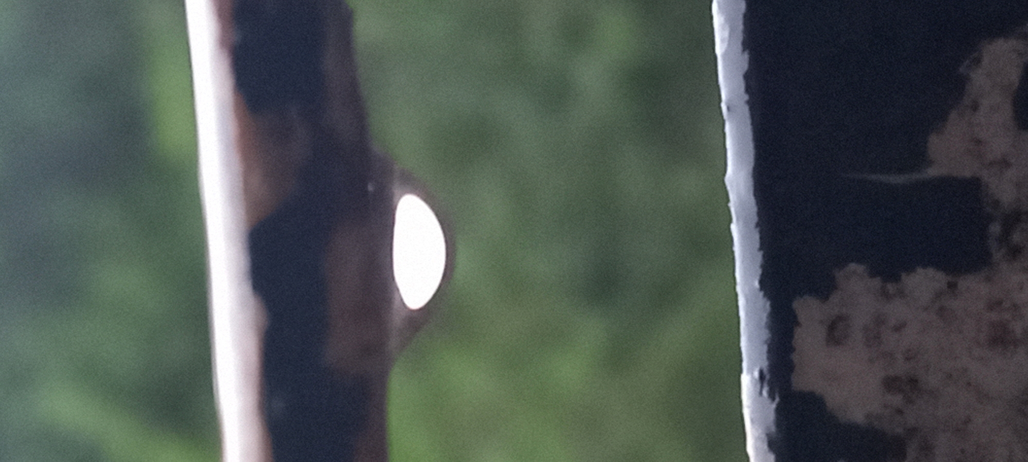 The image is a close-up shot of a water droplet hanging from a dark, weathered railing. The railing appears to be made of metal and shows signs of rust or peeling paint. The water droplet is illuminated from within, creating a bright, white oval shape. In the background, a blurred green landscape suggests an outdoor environment, possibly a park or garden. The lower portion of the image shows another dark railing with a textured, possibly concrete or stone surface. The scene is devoid of people and any specific indication of its location within Kryvyi Rih, Ukraine, beyond the assumption provided by the prompt.