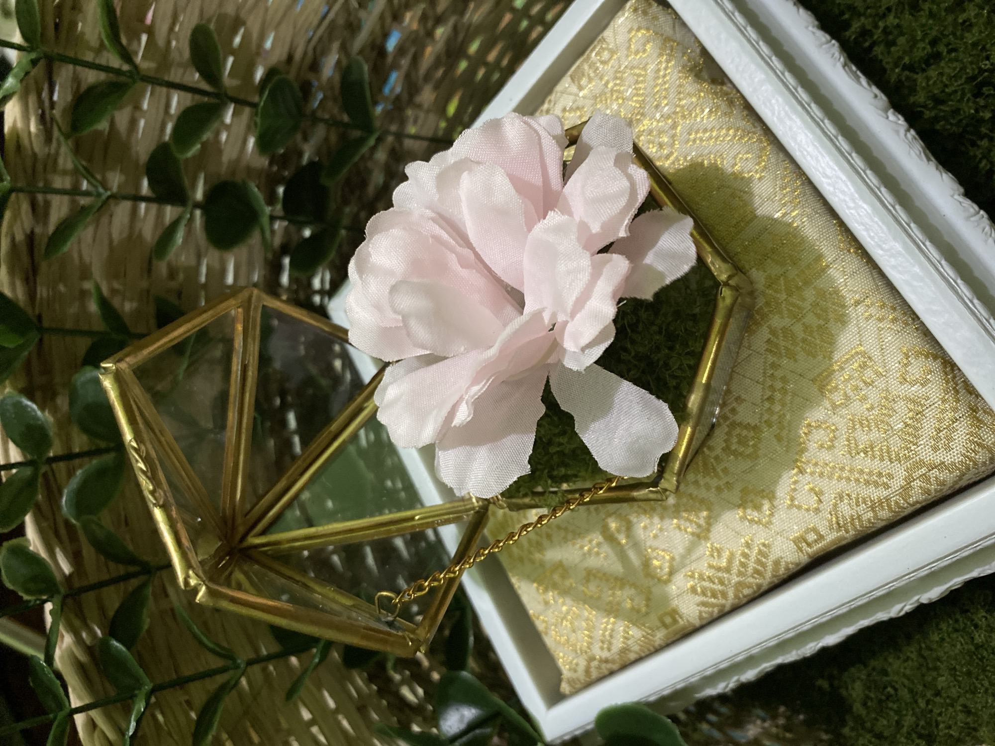 A close-up, overhead shot features a decorative ring box. The box is made of brass and glass, with a geometric, hexagonal shape. Inside the box, a bed of green moss supports a delicate pink fabric flower. A thin gold chain connects the lid of the box to its base.
The ring box rests on a white frame, which in turn is placed on a textured surface resembling woven straw. Behind the frame and box, more green moss is visible. A few artificial green leaves are scattered around the foreground and sides of the image. The overall scene evokes a sense of delicate presentation, possibly for a wedding or special occasion. The lighting appears soft and diffused, suggesting an indoor setting with controlled lighting. No text is visible in the image. The location context of Kuala Pembuang, Indonesia, does not provide specific visual cues in this image.
