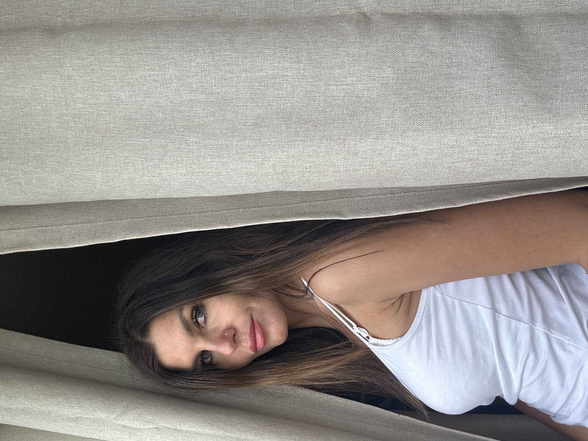 A woman is lying down, looking at the camera through a gap between two light gray surfaces, likely a curtain or blinds. She has long, brown hair and wears a white tank top. She has a nose ring on her left nostril. The background beyond the gap is dark. No evidence or information is present to indicate this picture was taken in Chaikovsky, Russia.