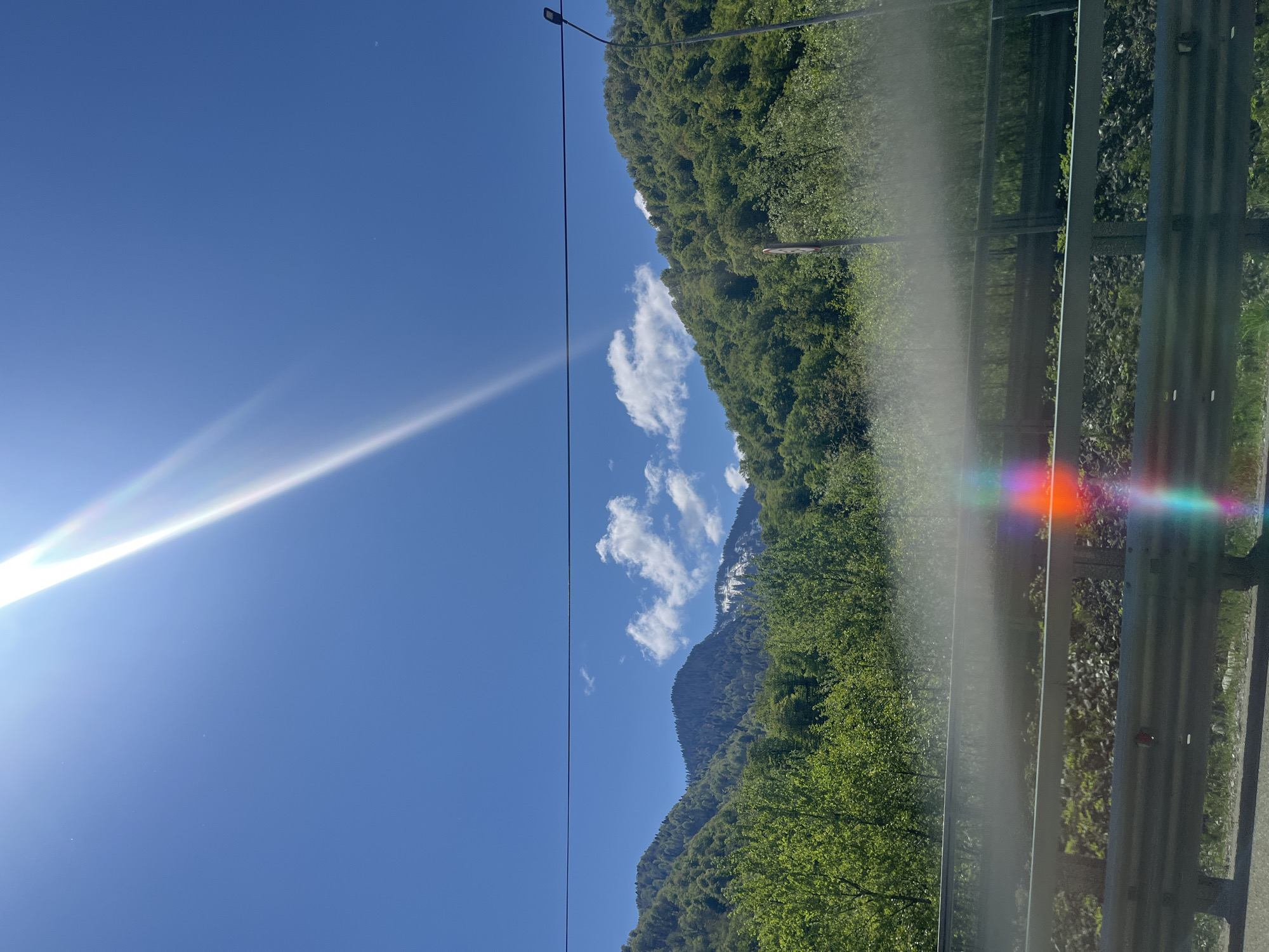 The image depicts a landscape with a clear blue sky and bright sunlight. A mountain covered in dense green forest is visible in the background, with some patches of white that could be snow or rock. There are a few fluffy white clouds in the sky near the mountain. A road with a metal guardrail runs along the right side of the image, indicating a roadside view. The sun's glare and lens flare are prominent in the sky and reflected on the guardrail. A power line is visible crossing the sky.