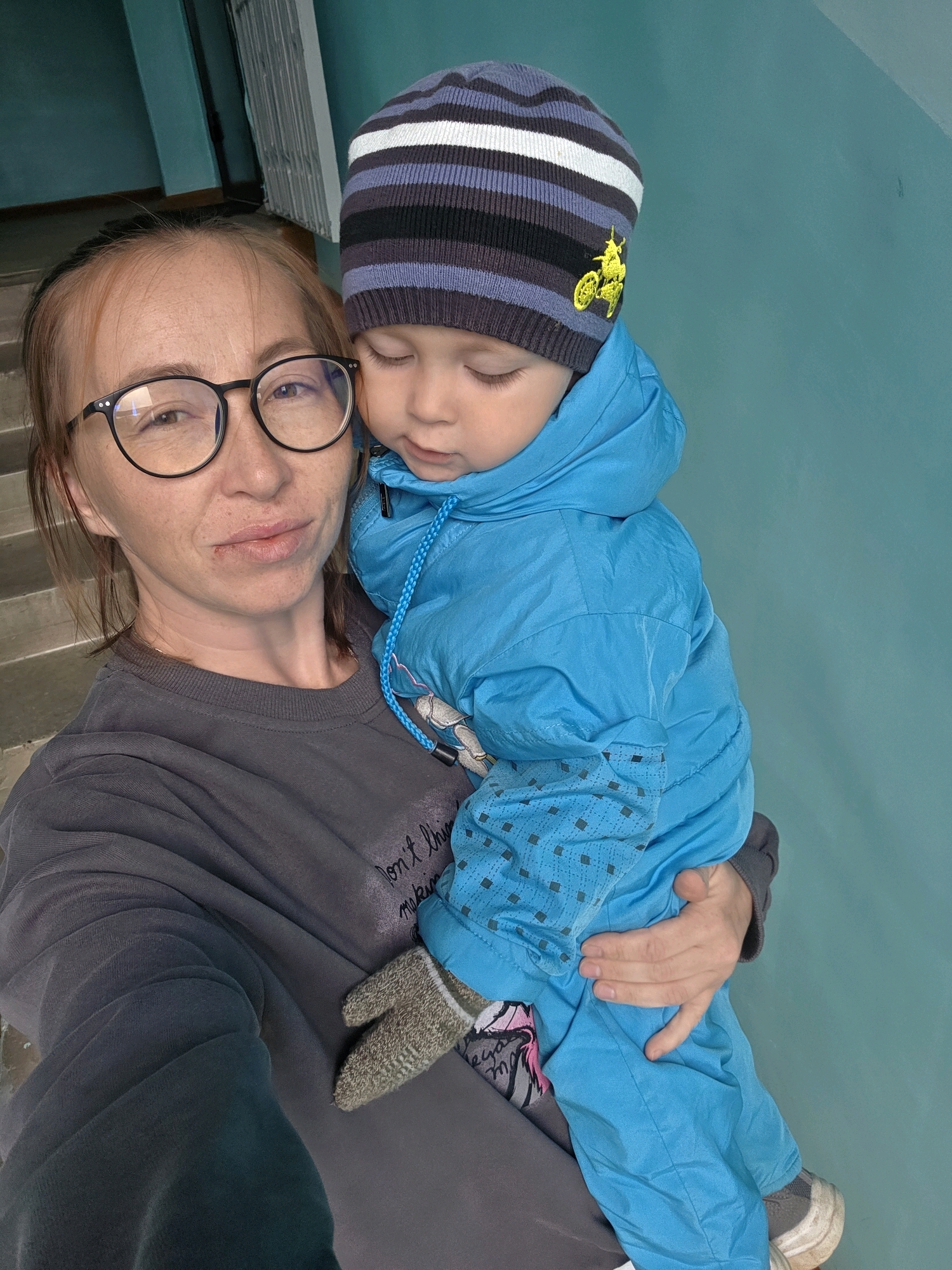 In a dimly lit indoor setting with teal-colored walls and a visible staircase, a woman with reddish-brown hair pulled back and wearing black-rimmed glasses holds a young child. The woman is wearing a dark gray sweatshirt. The child, who is being held by the woman, is wearing a blue hooded snowsuit and a striped beanie with a yellow motorcycle appliqué. The child's eyes are closed, and the woman is looking towards the camera. The scene appears to be in an apartment building or public institution, possibly in Chaikovsky, Russia, given the presence of what looks like a standard Soviet-era staircase.