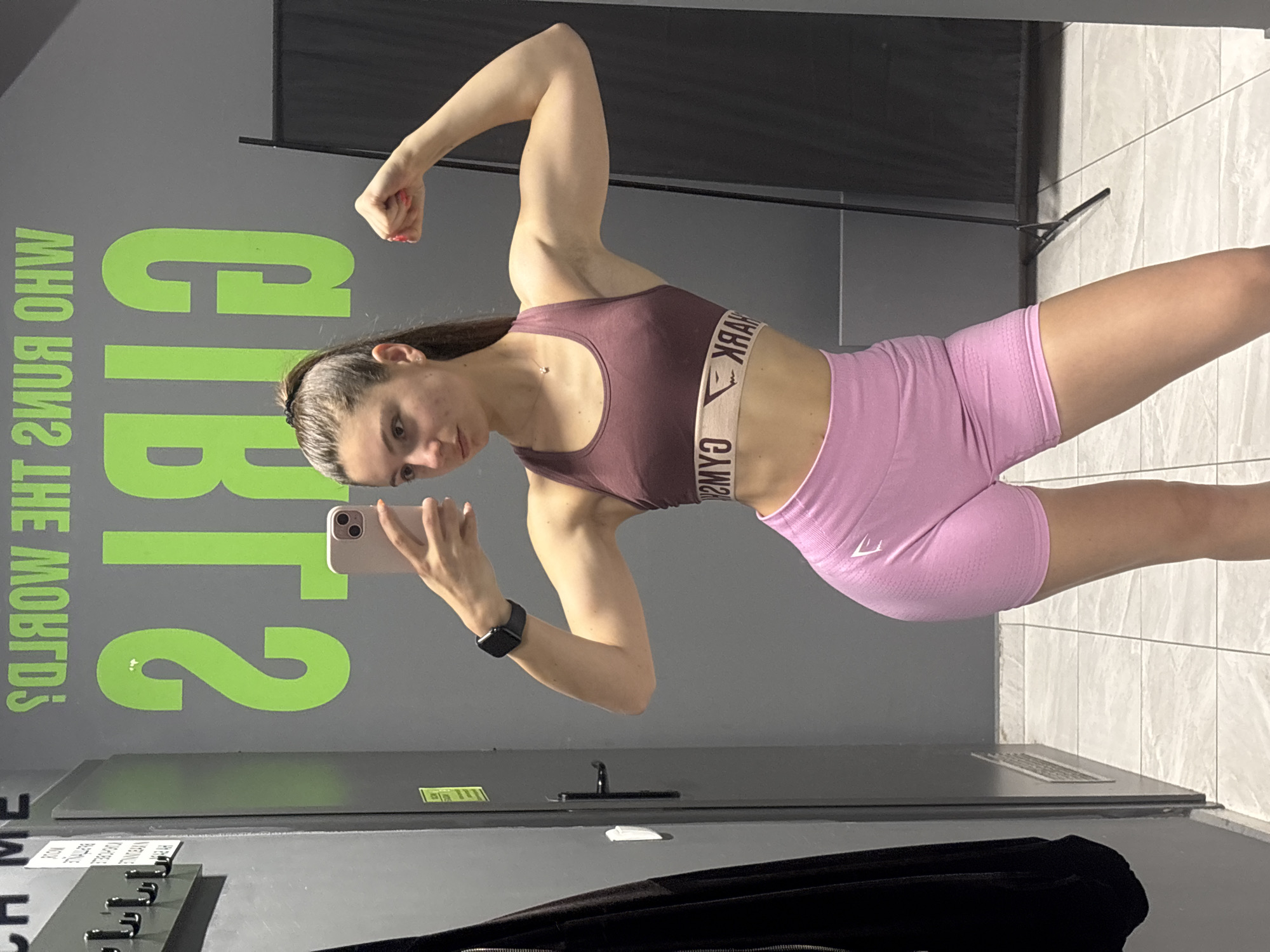 A young woman is taking a selfie in what appears to be a locker room or changing area, as evidenced by the hooks and lockers in the background. She is wearing a burgundy sports bra with a logo band around the bottom and light pink athletic shorts. She is flexing her left bicep while holding a phone in her right hand to take the picture. The wall behind her features a large green graphic that reads "GIRLS" and below that, "WHO RUNS THE WORLD?". The location details are not clear enough to determine the city of Varna, Bulgaria. The background includes a partition or partial wall covered in dark gray material, and to the right of that, a white tiled wall. A black coat is hanging on a hook.