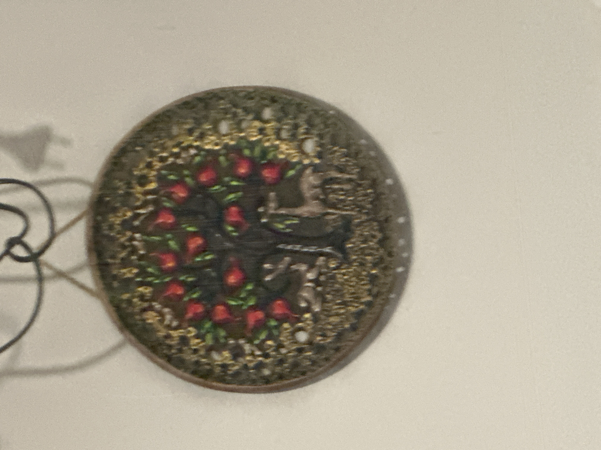 A slightly out-of-focus image depicts a round, decorative plate or plaque hanging on a light-colored wall. The plaque features a design with red floral elements, possibly roses or similar blossoms, arranged in a circular pattern around a central, darker shape that could be a tree or a stylized cross. The background of the plaque is heavily textured with gold and darker tones, creating an ornate or filigree-like effect. There is a dark, blurry object to the left of the plaque, possibly a lighting fixture. The exact location within Yerevan, Armenia, cannot be determined from the image.