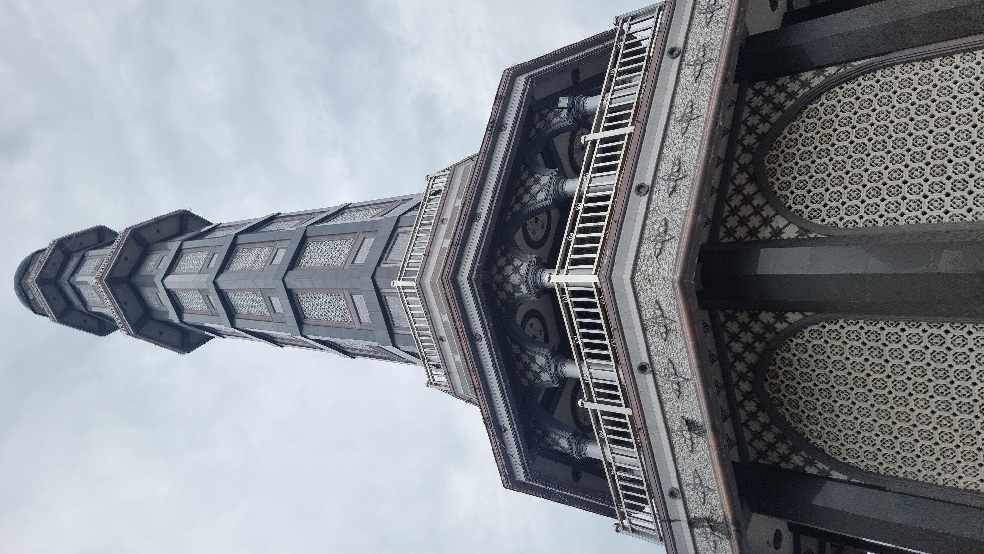 This is a low-angle, vertical shot of a minaret, likely belonging to a mosque, against an overcast sky. The minaret is tall and octagonal, constructed with dark grey stone panels featuring intricate latticework windows. The base of the minaret widens into a multi-tiered structure with balconies. These balconies are enclosed by white railings and supported by ornate columns with decorative carvings. The lower section of the structure has large arched openings filled with a repeating geometric pattern of light-colored stone. The overall architectural style is modern with traditional Islamic influences. The location is identified as Keudekruenggeukueh, Indonesia. There are no people or visible actions in the image.