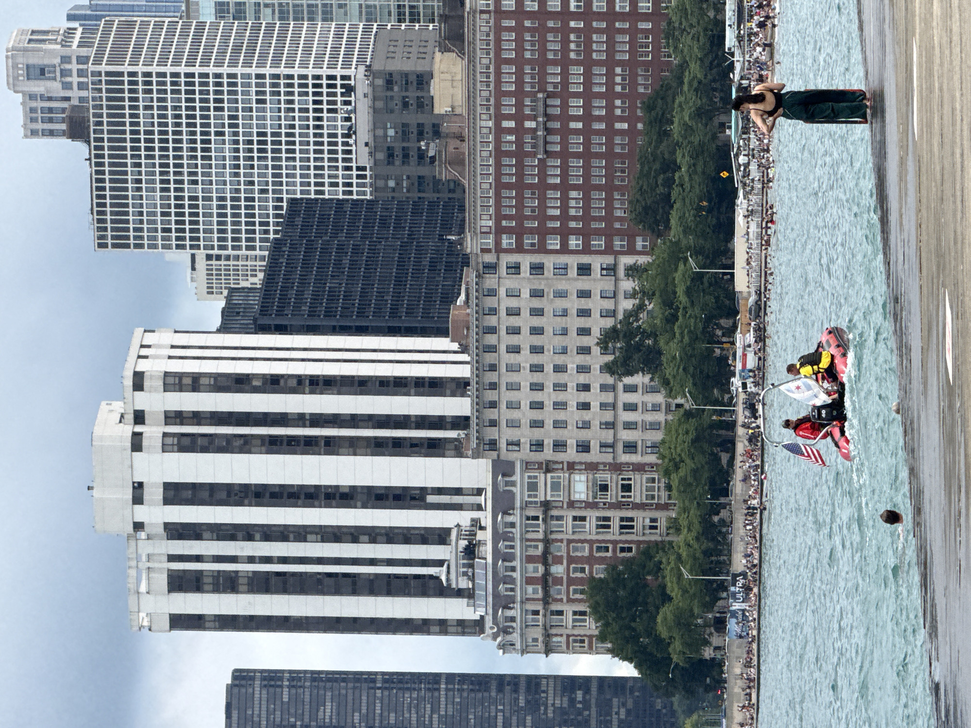 The image captures a vibrant scene along the Chicago lakefront. In the foreground, a person stands on a concrete pier, looking out at the water. The water is choppy, with several people swimming and a small boat with two individuals, one wearing a red jacket and the other a yellow life vest, navigating the waves. The boat is adorned with American and Chicago flags. A crowd of people can be seen gathered on the shore in the background, with various tents and structures suggesting a public event or beach area. Looming behind the shoreline is a dense cluster of iconic Chicago skyscrapers, including the striking black and white patterned building, the red brick apartment buildings, and other high-rise structures that define the city's famous skyline. The sky is overcast, contributing to a slightly muted but still lively atmosphere. The overall impression is one of a bustling urban waterfront on a cloudy day.