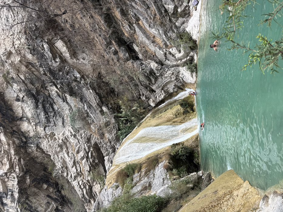 The image depicts a natural outdoor scene in San Miguel Atlapulco, Mexico, dominated by a large waterfall cascading over multi-tiered, yellowish-brown rock formations into a prominent turquoise-colored pool. Steep, stratified rock cliffs, varying in shades of grey, black, and light beige, rise on the right and in the background, exhibiting sparse, dry vegetation and some small green plants. The water in the pool appears clear and calm.
Four individuals are present within the turquoise pool. In the right-center, a male with dark hair is submerged chest-deep, facing towards the left. Near the base of the main waterfall, a person wearing an orange flotation device is floating on their back. Further to the right, at a smaller cascade where water enters the pool, two additional individuals are visible: one person in a red top is wading or sitting directly beneath the descending water, and a second person is partially obscured behind them. Green leafy foliage is present in the immediate lower right foreground, partially obscuring the water's edge.