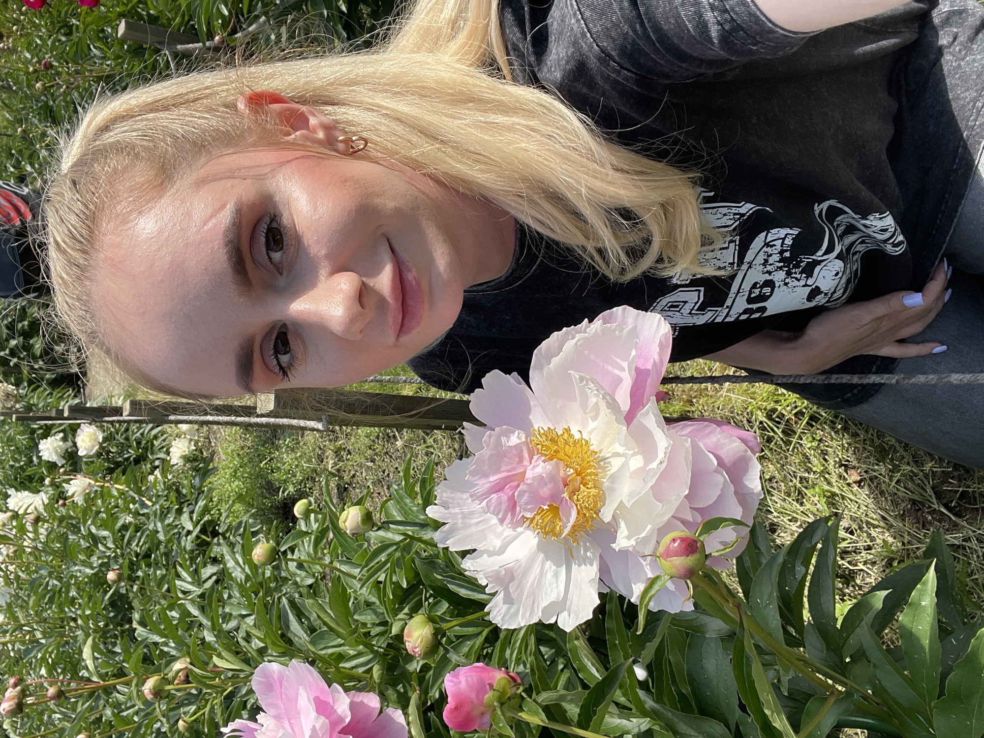 A young woman with blonde hair tied in a ponytail is captured in a close-up, selfie-style shot. She is wearing a dark t-shirt with a graphic print and light-colored jeans. Her left hand rests on her lap, and her right arm is extended to hold the camera. The woman has light makeup, including pink blush and eyeshadow, and wears a small hoop earring in her visible left ear.
She is positioned outdoors, surrounded by a garden filled with pink and white peonies. Several large peony blooms are in focus, showcasing their layered petals and yellow centers. Other flowers and green foliage form the background, with hints of wooden stakes and grass visible. The lighting suggests it is daytime with natural sunlight. The image does not contain any specific visual cues that would allow for the identification of its location in Vladivostok, Russia.