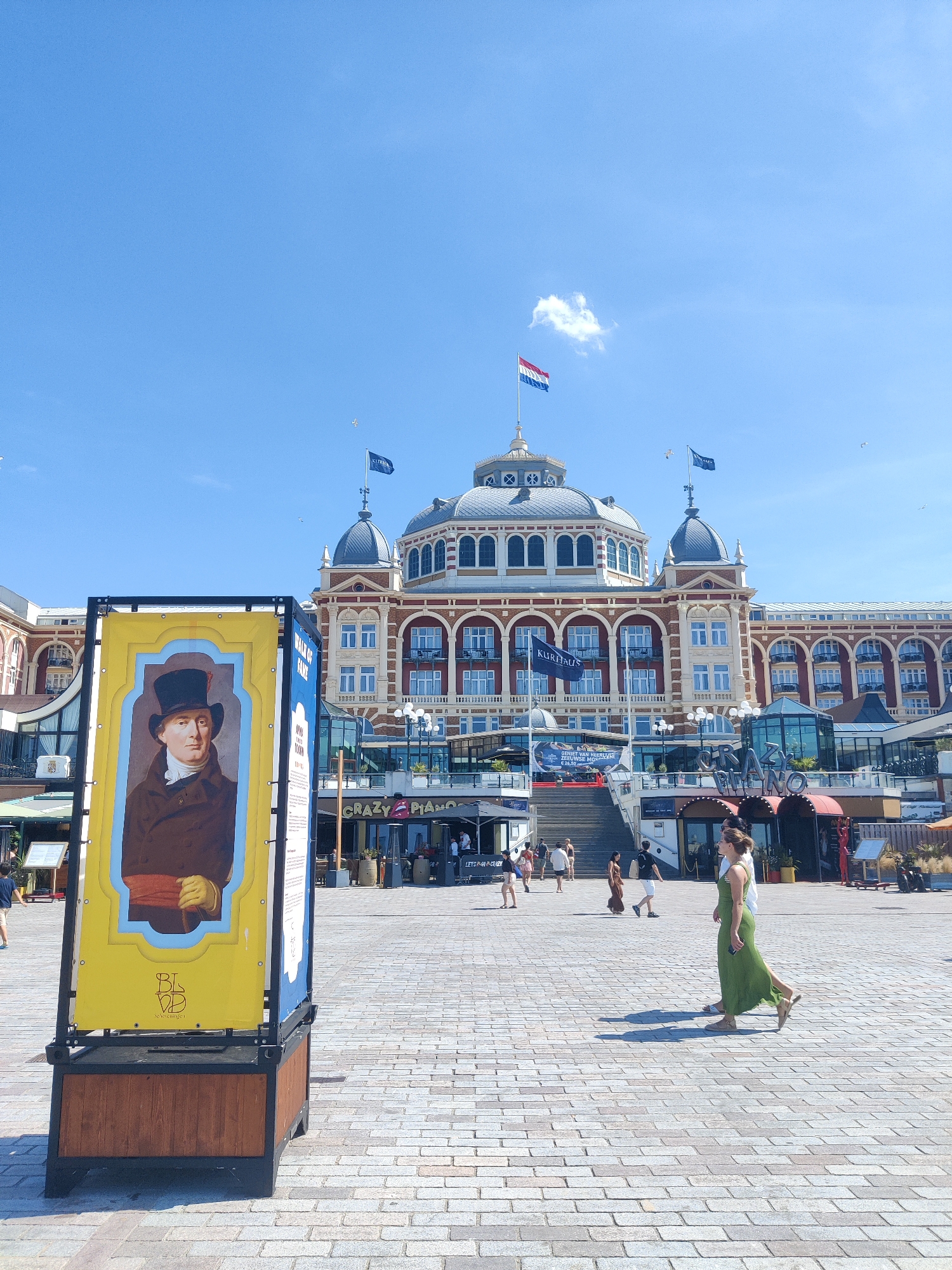 The image depicts a sunny day in The Hague, Netherlands, specifically in front of the Kurhaus hotel. The Kurhaus is a large, historic hotel with distinct red brick and domed architecture, flying the Dutch flag from its central dome. In the foreground, a large, yellow display board features a portrait of a man in a top hat and waistcoat, identified as part of a "Walk of Fame." The ground is paved with cobblestones. Several people are visible walking in the plaza, including a woman in a green dress and a man in a white shirt. Various establishments, including "Crazy Piano," are visible on the lower levels of the Kurhaus building. The sky is clear blue with a few scattered clouds and birds.