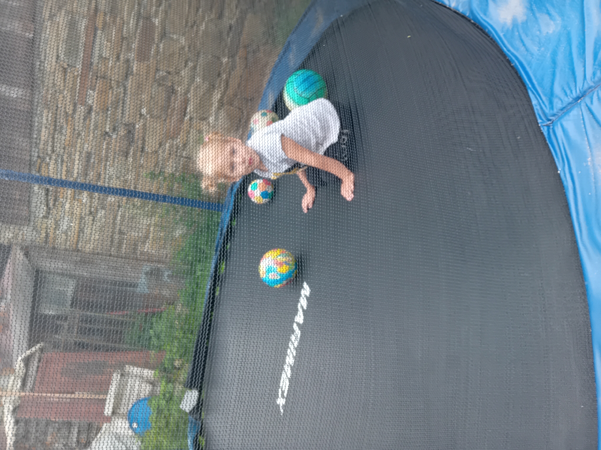 A young girl with blonde hair in two pigtails is sitting on a trampoline. She is wearing a white t-shirt with a pattern and black shorts. The girl is looking towards the camera. There are three balls on the trampoline with her: one multicolored ball, one patterned ball, and one green and blue ball. The trampoline is dark grey and has a blue edge. In the background, a brick wall and a wooden structure are visible, possibly part of a house or shed. The scene appears to be outdoors, in a yard. The image was taken in Zuberec, Slovakia.