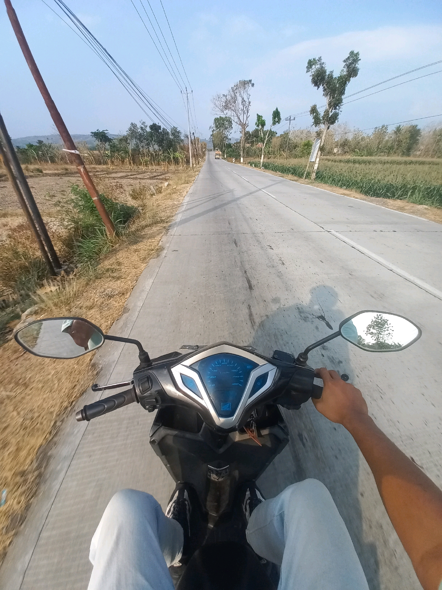 The image shows a first-person perspective of someone riding a motorcycle on a paved road in Baturetno, Indonesia. The road is a two-lane street with a white dividing line. On the left side of the road, there is dry grass and dirt, with a wooden pole and power lines running along it. On the right side of the road, there is a field of green crops, likely corn. Trees are visible in the distance, with a clear blue sky overhead. The motorcycle's dashboard is visible, showing a speedometer and other gauges. The rider's hands are on the handlebars, and their legs are visible in the lower part of the frame. A shadow of the rider is cast on the road. A yellow vehicle is visible in the distance on the road.