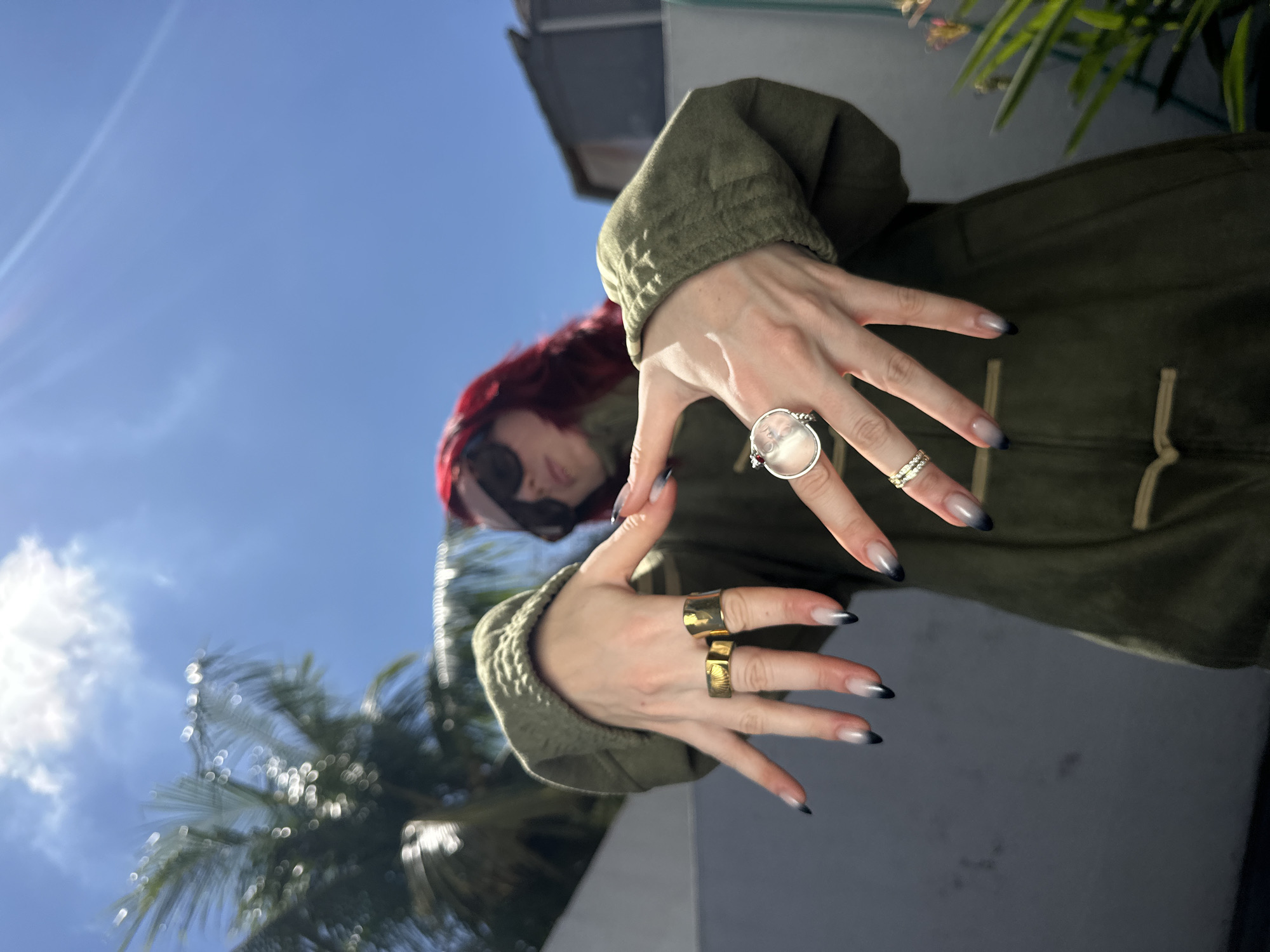The image shows a person with bright red hair, wearing a green jacket and sunglasses. They are standing outdoors with their hands outstretched, showing off several rings and manicured nails. In the background, palm trees and a blue sky with a few clouds are visible, suggesting a tropical or warm climate. The person's pose and expression seem confident and possibly playful. The overall scene appears to be a casual outdoor setting, potentially in a city like North Kuta, Indonesia, given the presence of palm trees. The lighting indicates a sunny day.