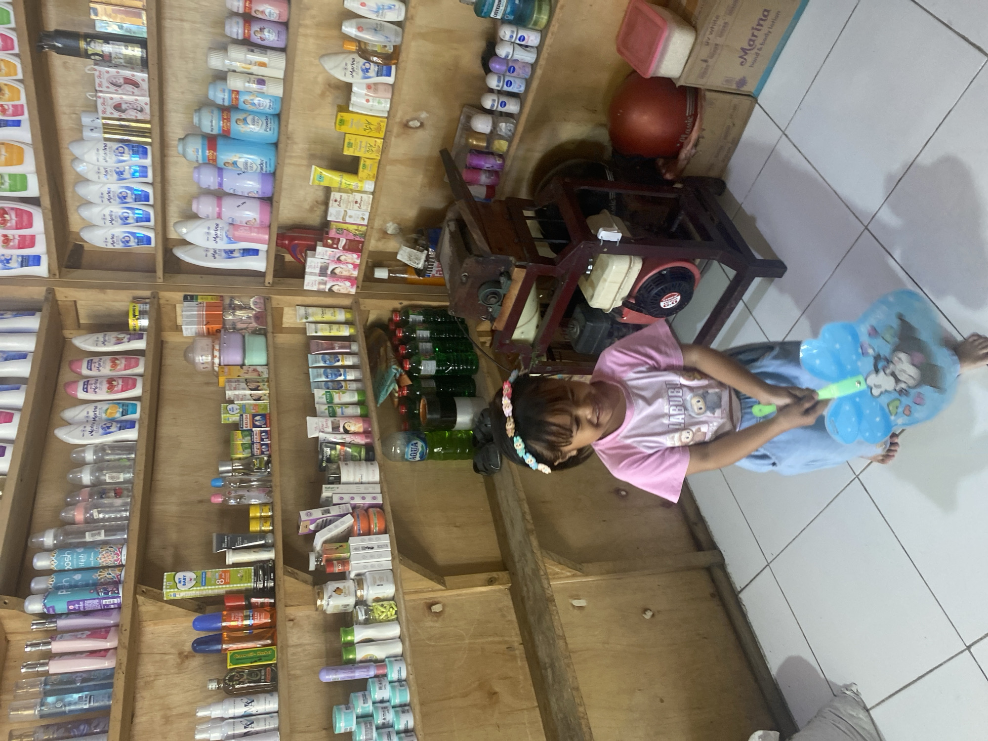 The image shows a young girl holding a blue butterfly-shaped balloon in a small shop in Ta', Indonesia. The shop is filled with shelves stocked with various personal care products and household items. The girl is wearing a pink t-shirt with cartoon characters and blue pants. She is smiling and looking at the balloon. The shop appears to be a local convenience store or a small pharmacy, with products like lotions, soaps, and detergents displayed on wooden shelves. A small engine-powered machine, possibly used for some sort of processing or manufacturing, is visible behind the girl. The overall impression is of a modest retail environment catering to local needs.