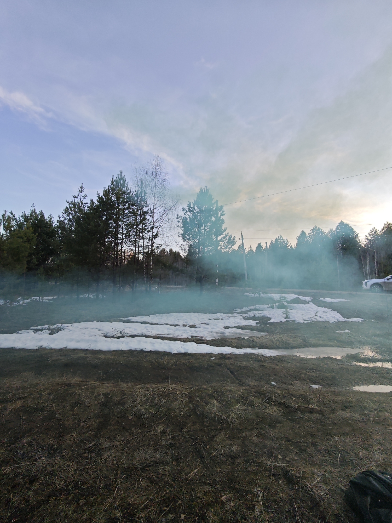 An outdoor scene captures a landscape in Dubovaya, Russia, characterized by the transition from winter to spring. The foreground consists of rough, dry, brown ground and sparse vegetation, with several patches of melting snow and small puddles of water reflecting the sky.
A prominent plume of teal-blue colored smoke billows across the mid-ground, partially obscuring a dense line of trees that forms the backdrop. These trees are a mix of tall pine or spruce evergreens and bare deciduous trees, suggesting early spring before leaf-out. Through the smoke, the darker silhouettes of the tree trunks are visible.
In the upper portion of the image, the sky is partly cloudy with a pale blue hue, transitioning to lighter and warmer tones towards the right horizon, indicative of either late afternoon or early evening. A utility pole with power lines is visible on the right side, extending horizontally across the frame. A white car is partially visible on the far right, parked behind the smoke and near the tree line. No people are visible in the image. In the bottom right corner, a dark object, possibly a bag or backpack, rests on the ground. The overall atmosphere is somewhat tranquil yet made dynamic by the spreading smoke.