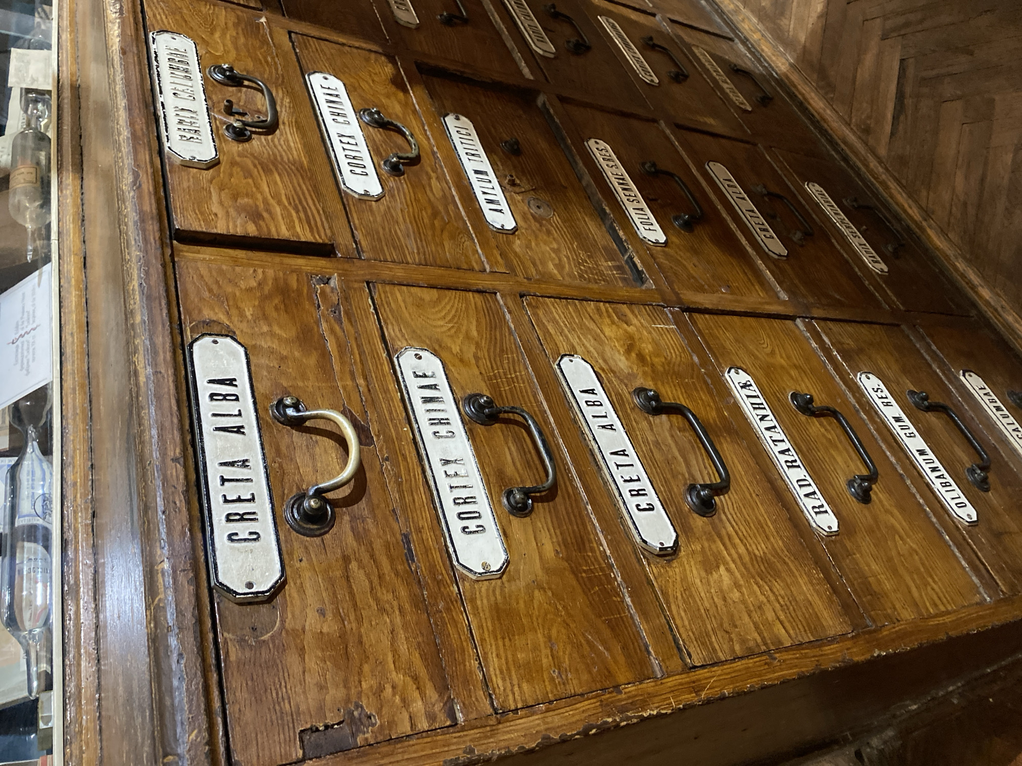 The image shows a close-up of an antique wooden apothecary cabinet with multiple drawers, each labeled with the contents in Latin. The cabinet is made of dark wood with brass handles and is situated in what appears to be a museum or historical pharmacy. The labels visible include "CRETA ALBA," "CORTEX CHINAE," "RADIX CALUMBAE," "AMYLUM TRITICI," "FOLIA SENNAE S.RES.," "RADIX RATA NHIA," "OLIBANUM GUM. RES.," and "RADIX SERPENTARIAE." The cabinet is well-maintained, showcasing its historical significance. This cabinet is located at the Museum of Pharmacy in Lviv, Ukraine.