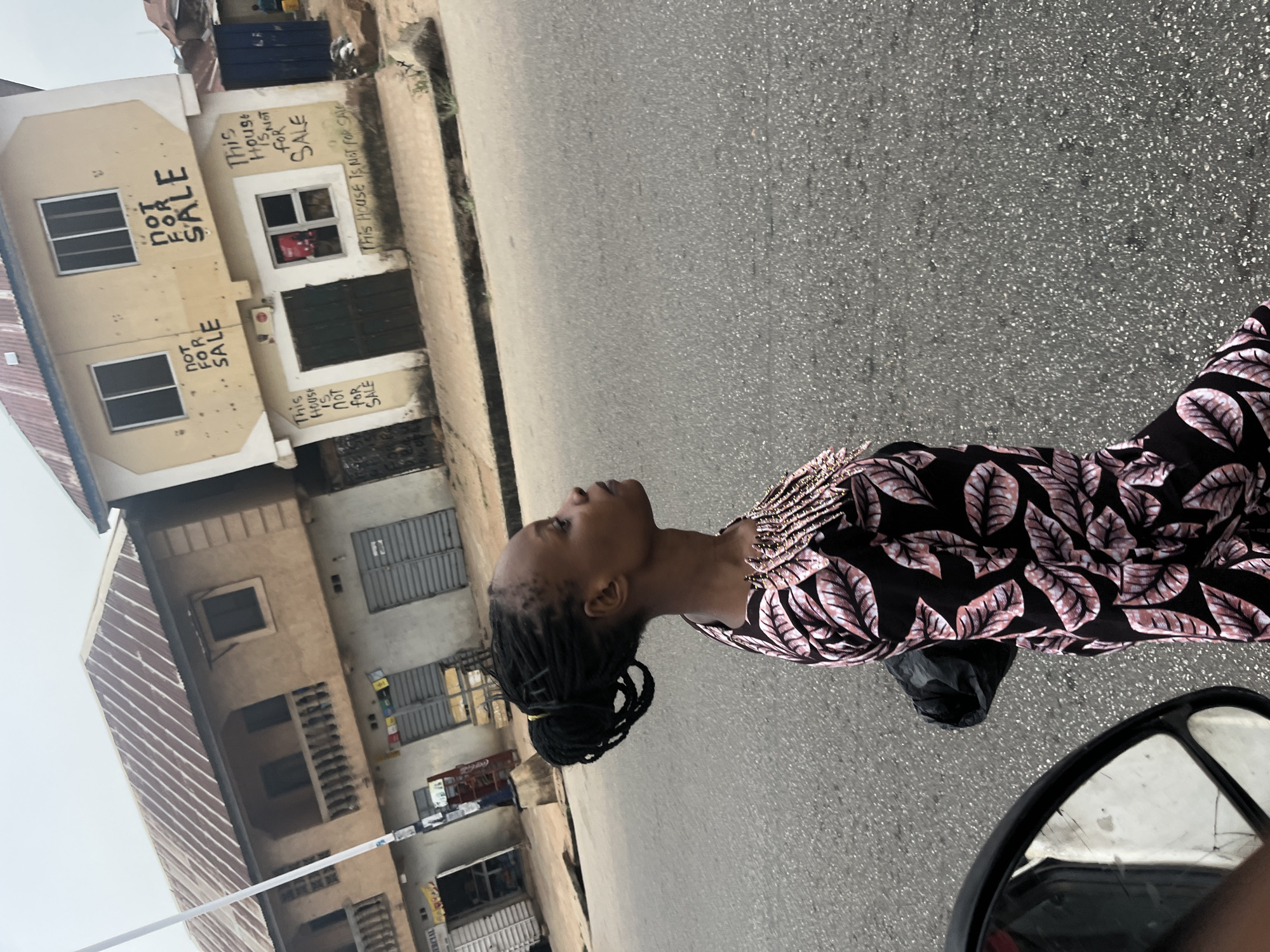 A woman with dark braided hair and a black floral dress is standing on a gray asphalt road, looking upwards and to the left. Behind her are buildings, one of which is marked with graffiti stating "This House is NOT FOR SALE." The location is identified as Jos, Nigeria.