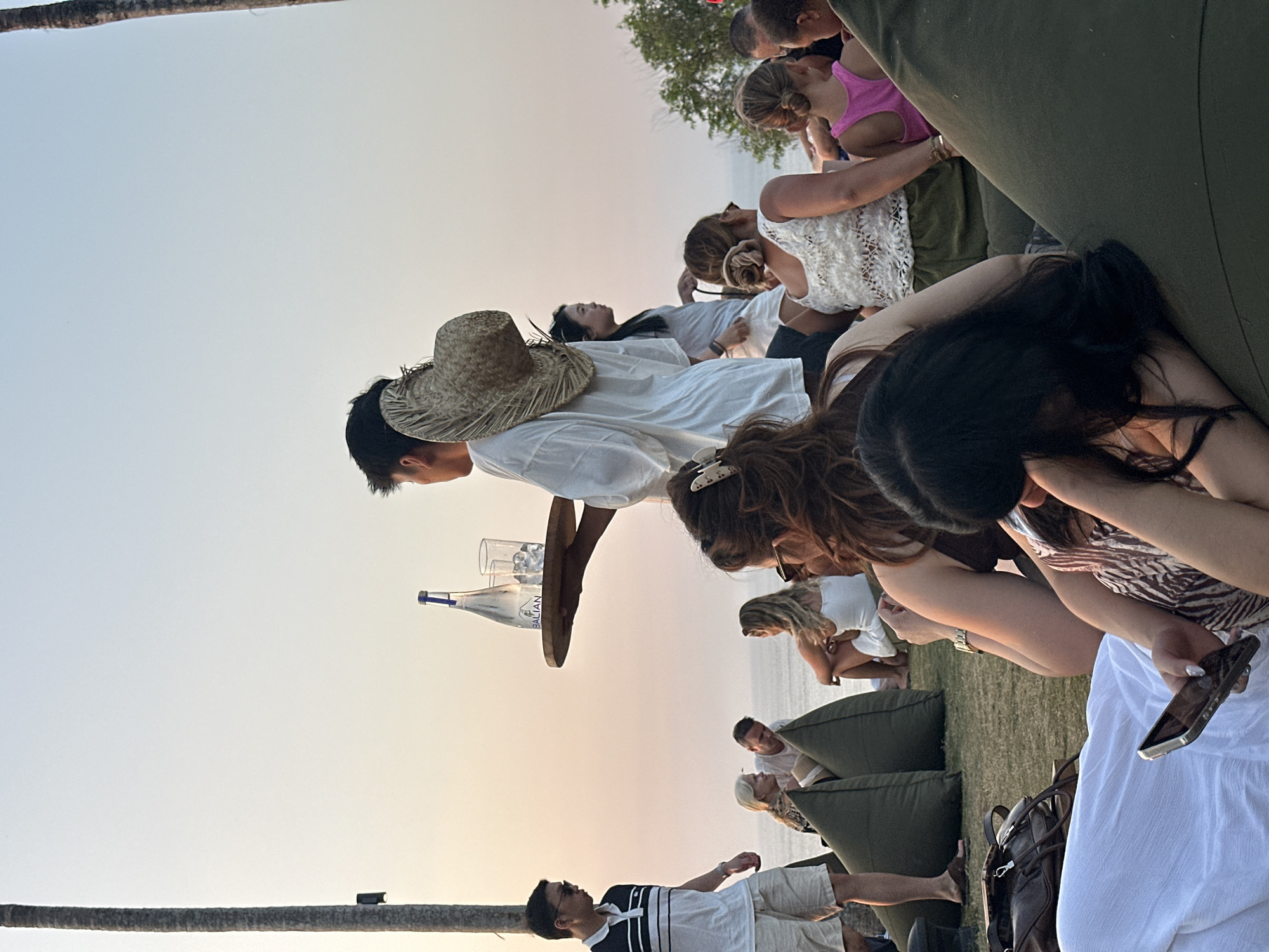 This is a medium shot image taken at sunset in Kuta Selatan, Indonesia. The scene depicts a group of people relaxing on beanbag chairs on a grassy area overlooking the ocean. A server, wearing a white shirt and a straw hat, is carrying a tray with a bottle of water and two glasses of ice. In the foreground, a woman is holding a smartphone. The sky is a soft orange and pink, indicative of a sunset. The overall atmosphere appears relaxed and social.