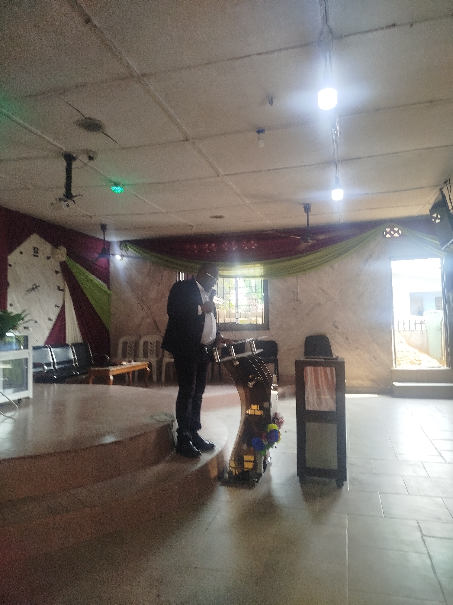 The image shows the interior of a building, potentially a church, located in Ikot Mbonde, Nigeria. A man dressed in a dark suit and a light-colored shirt is standing behind a lectern, facing forward. The lectern has a decorative front with flower arrangements. To the right of the lectern is a small dark structure with a recessed panel. The background features a raised platform with chairs and a small table. A large clock-like decoration is on the left wall, and fabric drapes adorn the walls behind the platform. A window with security bars is behind the lectern. There's an open doorway with a bright outdoor scene visible, leading to a fenced area. The ceiling is tiled, with visible light fixtures and a ceiling fan. The floor is tiled in a light color.