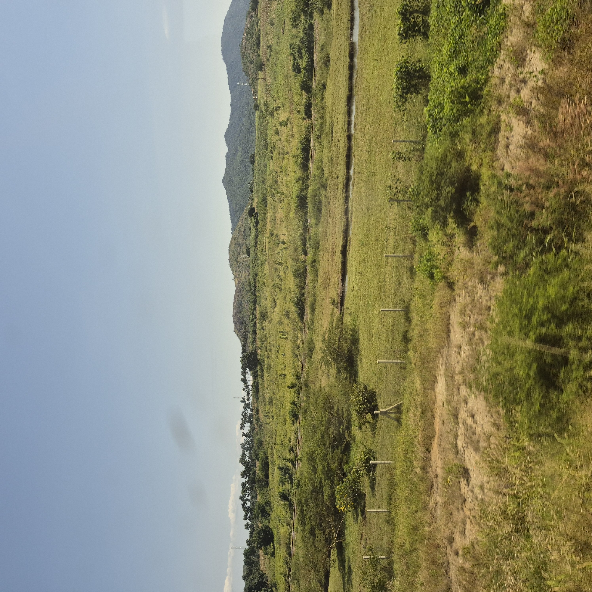 The image depicts a rural landscape in Mkambarani, Tanzania, under a clear blue sky with a few scattered clouds. In the foreground, a dry, grassy area is demarcated by simple fence posts. Beyond this, a gently sloping green field stretches towards a treeline. A narrow, dark body of water or a dry riverbed runs horizontally across the mid-ground, separating this field from another expanse of green vegetation. Further in the distance, rolling hills covered in dense foliage rise to meet the horizon. The vegetation appears lush and green, suggesting a tropical or subtropical environment. The overall impression is one of a tranquil, undeveloped natural setting.