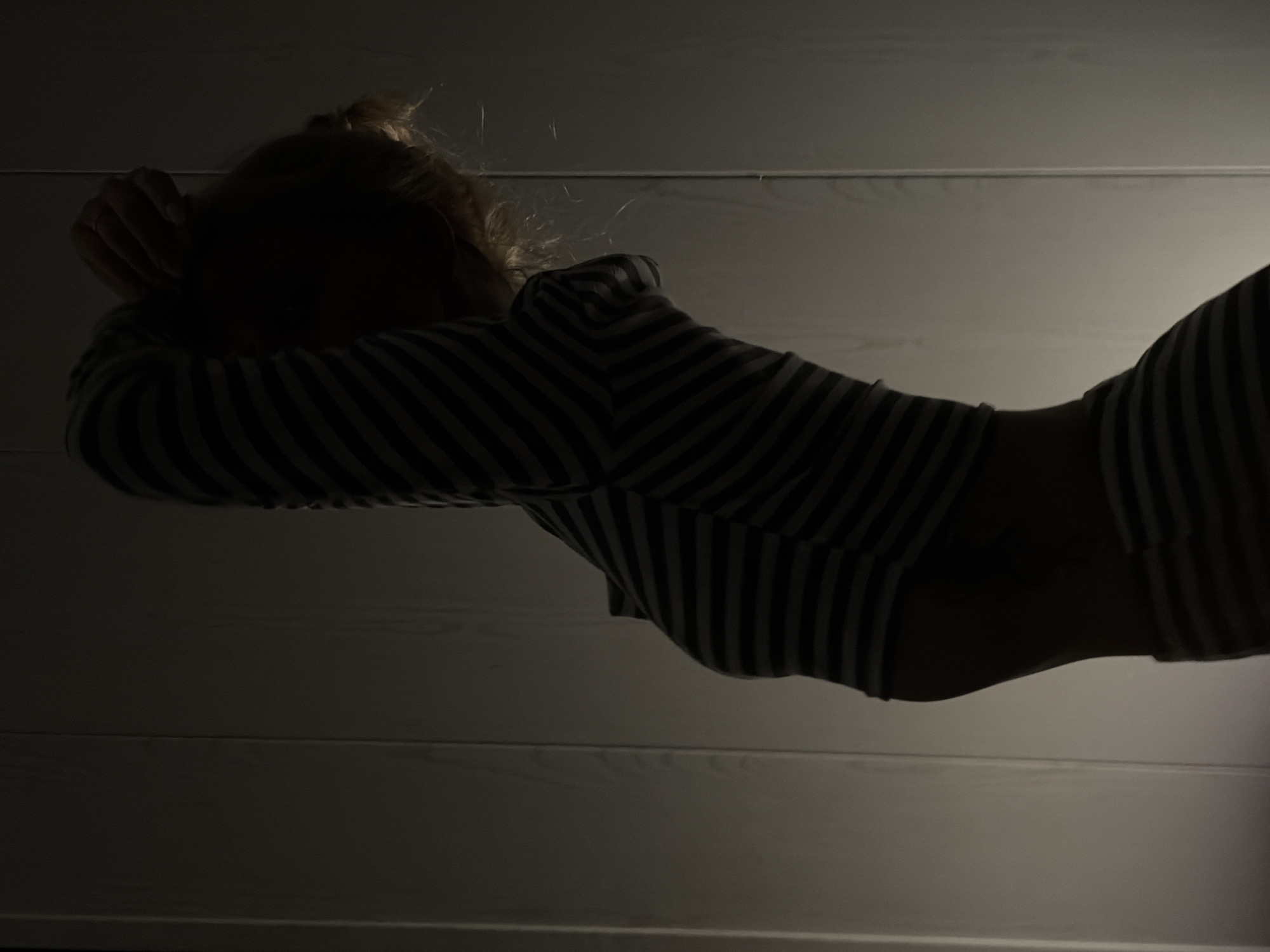 A low-angle shot depicts a person lying down in a dark room. The person has fair hair gathered on their head and is wearing a striped shirt with horizontal black and white stripes. Their hands are positioned near their head. The background consists of a wall with horizontal planks, which are faintly illuminated. The image is overall dimly lit with shadows obscuring details. There is no indication of the location being Chaikovsky, Russia.