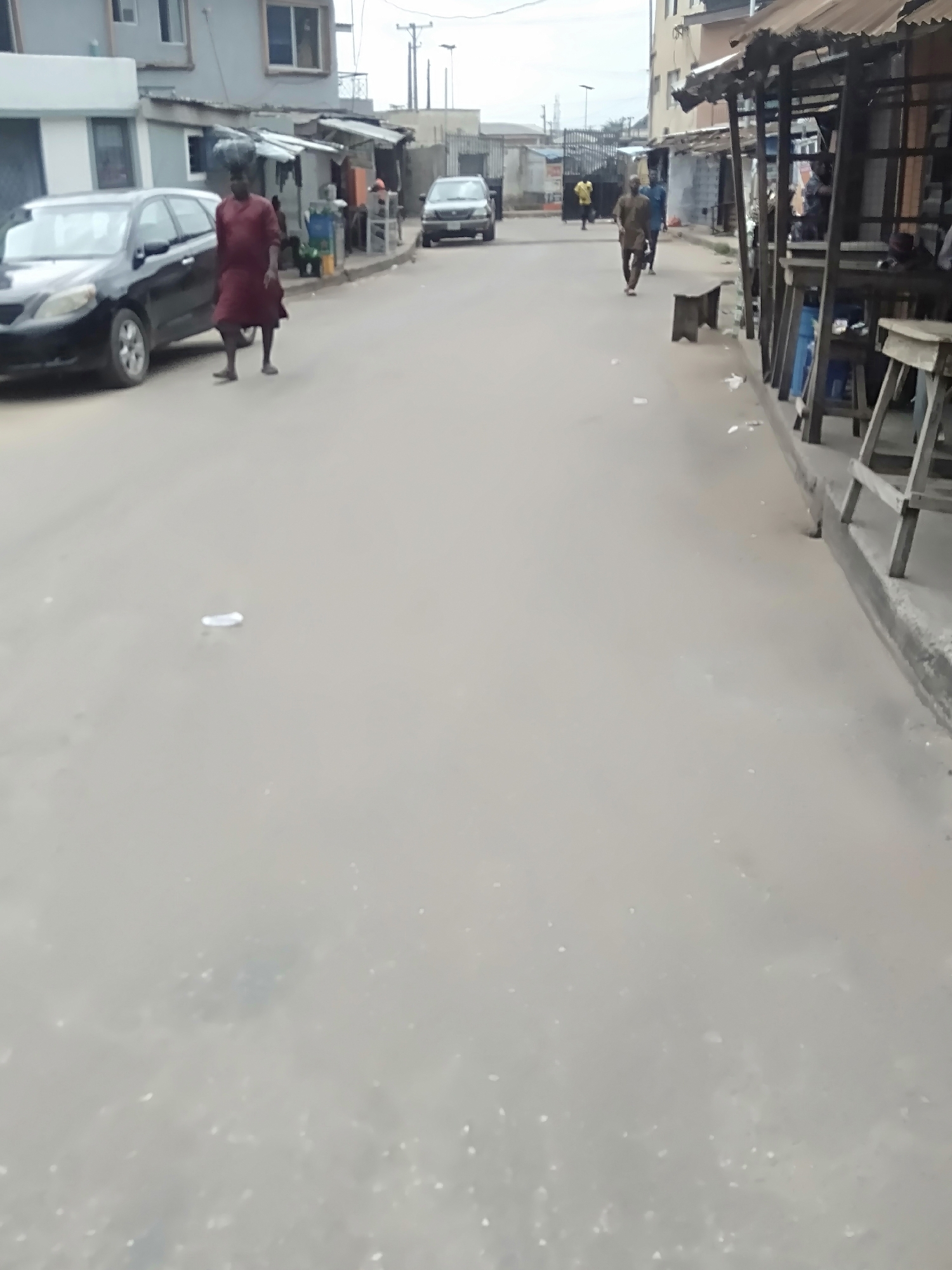 The image shows a street in Shangisha, Nigeria. A black car is parked on the left side of the street. A person is walking in front of the car, carrying something on their head. Further down the street, a person is walking away from the camera. A building is on the right side of the street, partially covered by structures that look like shops. There is litter scattered on the street.