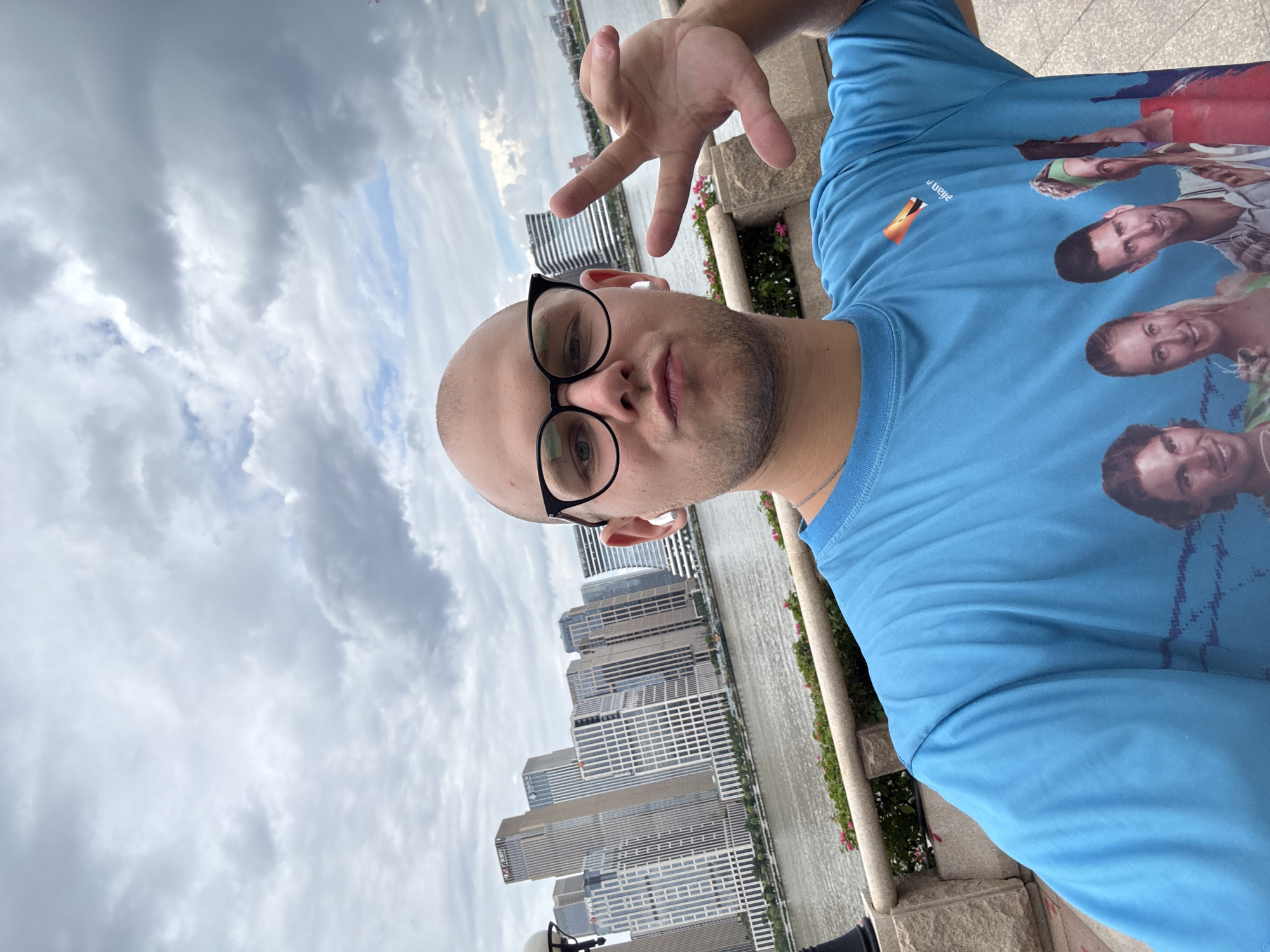 This is a vertical, slightly blurred selfie of a bald man wearing glasses and a blue t-shirt. He is positioned in the foreground, with his face and upper body dominating the frame. The man is looking directly at the camera with a neutral expression. He has a few days' growth of stubble on his face. He is wearing black-rimmed glasses and appears to have white earbuds in his ears. His right hand is raised in the frame, making a peace sign.
The man is wearing a bright blue t-shirt that has a graphic print of several people's faces on it, seemingly a group of tennis players. Some text is visible on the t-shirt, partially obscured. The text "Julian" is visible above a small orange and black logo on the left chest area of the shirt.
In the background, a river or body of water stretches horizontally across the image. Beyond the water, a cityscape with numerous tall buildings is visible. The buildings are modern and appear to be office or apartment blocks. The sky is overcast with a mix of grey and white clouds, suggesting it might be daytime but not particularly sunny.
The setting appears to be an outdoor promenade or walkway, as indicated by the stone railing and planters with pink flowers to the right of the man. The overall impression is a casual snapshot taken outdoors with a city backdrop. The rotation of the image suggests it was taken with the camera held horizontally, then rotated for the vertical orientation.