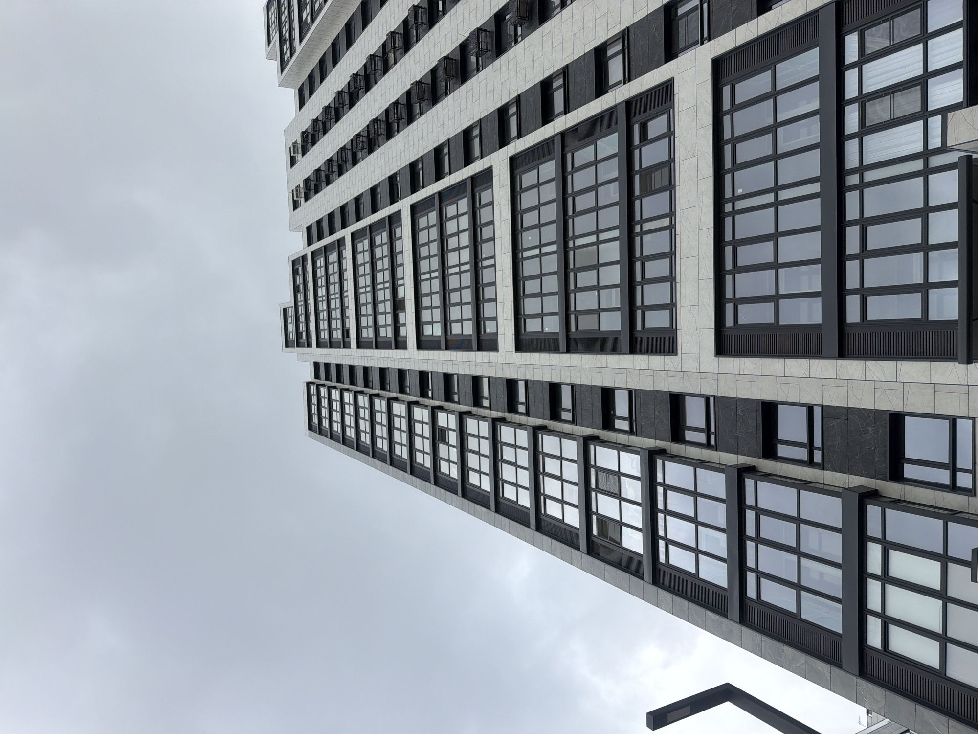 The image shows a tall, modern building in Rostov-on-Don, Russia, viewed from a low angle. The building features a facade with a grid-like pattern of windows and contrasting light-colored stone or tile. The windows are large and have black frames. The sky is overcast and gray. A portion of a street light fixture is visible in the lower portion of the image.