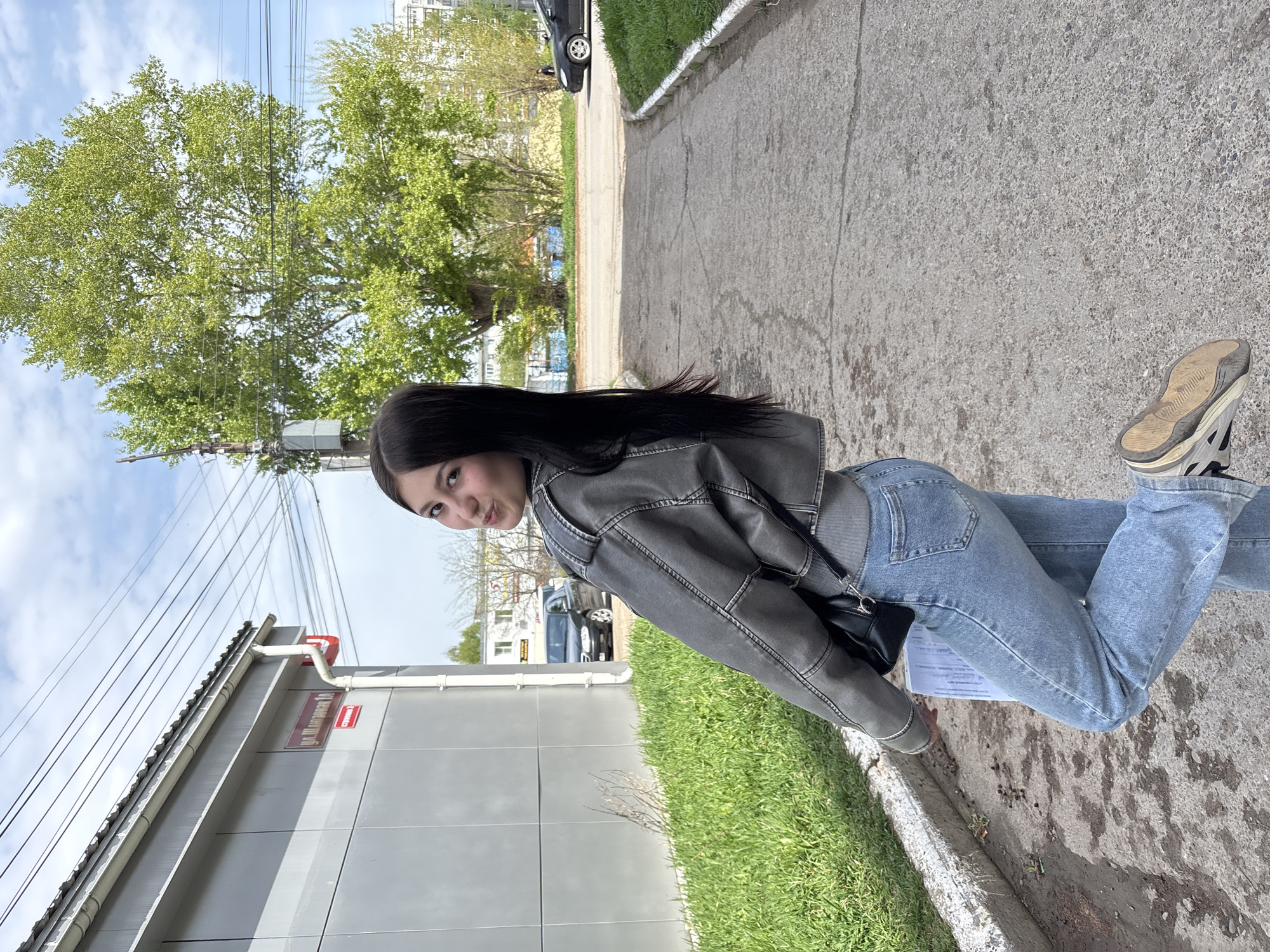 A young woman with long, dark hair stands on a sidewalk next to a building in Chaikovsky, Russia. She is wearing a gray jacket and blue jeans, and she has a black purse slung over her shoulder. She is holding a paper in her hand. A tree and a grassy area are in the background. The building has a sign in Russian that says "ул. Шаманская 10". Electrical wires are visible above the building.