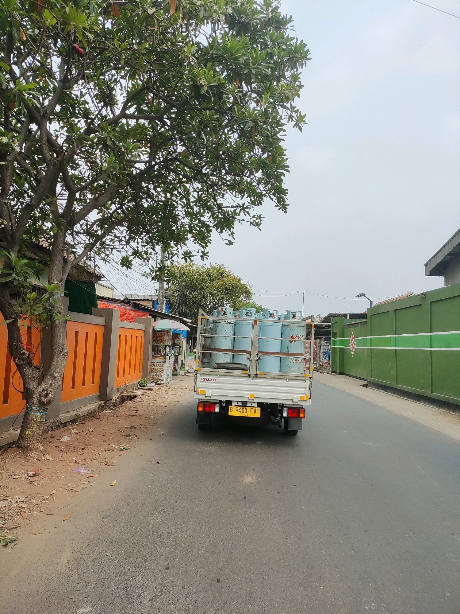 The image depicts a street scene in Wanajaya, Indonesia, with a white Isuzu truck carrying several large blue gas cylinders driving away from the camera. The truck's license plate reads "B 9401 FVT". To the left of the road is an orange fence with a textured pattern, bordered by a dirt path with some debris. A tree with green leaves and a visible trunk is positioned in the foreground, close to the fence. Behind the fence, several buildings are partially visible, including one with a green awning and another with a red tarp. In the background, a large green wall with white horizontal stripes runs along the right side of the road. The sky is overcast.