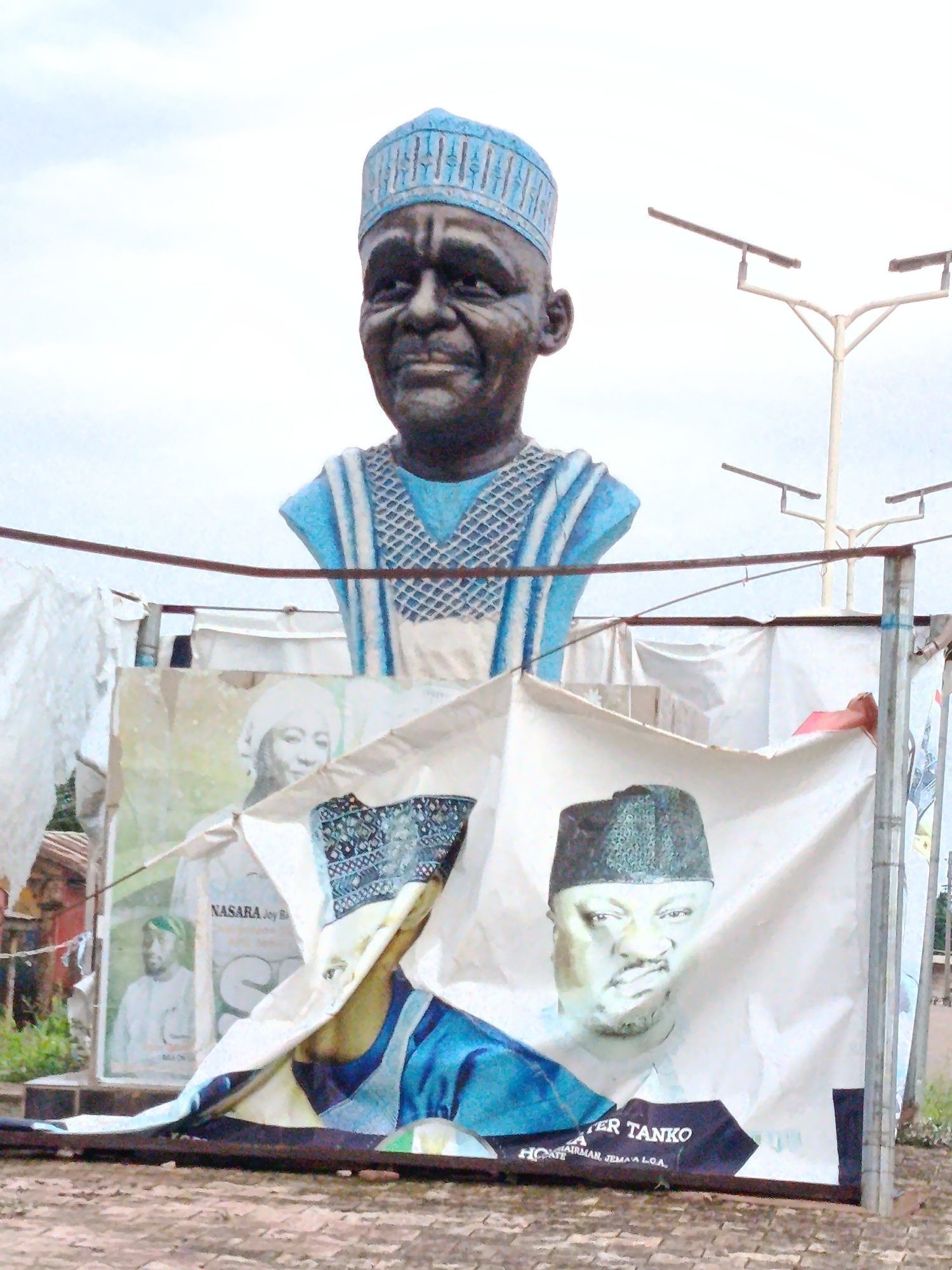 This image depicts an outdoor scene in Maigizo, Nigeria, dominated by a large, blue-hued bust of a man wearing a patterned cap and a traditional outfit. The bust appears to be made of concrete or a similar material. In front of the bust, and partially obscuring it, are several large banners, also featuring portraits of men in traditional attire and caps. One banner prominently displays the text "NASARA JOY B" and a portrait of a man with a white cap. Another banner features a portrait of a man with a black cap and the text "TER TANKO CHAIRMAN JEMA L.O.A.". These banners are hung on a makeshift structure, likely for an event or political campaign. The background shows a cloudy sky and some greenery, suggesting a warm climate. The ground in the foreground is a reddish-brown, brick-like surface. The overall impression is of a public display, possibly related to a local celebration or political gathering in Maigizo.