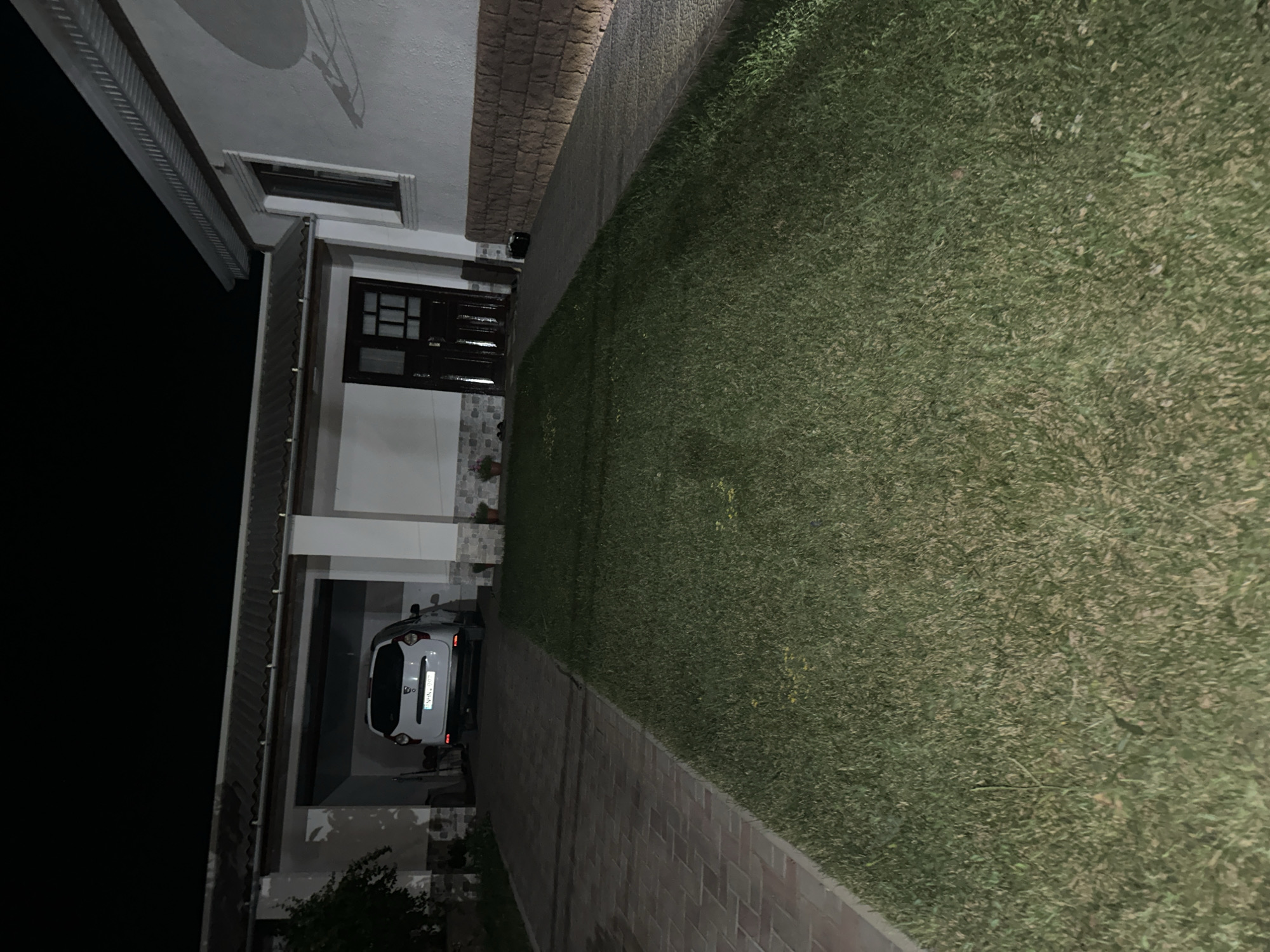 The image depicts the exterior of a house at night in the city of Bolohan, Moldova. A white Dacia Duster is parked under a covered carport, with its rear facing the camera. The house has a light-colored facade, a dark brown door with glass panes, and a window with a dark frame. A paved driveway made of interlocking bricks leads to the carport. Adjacent to the driveway is a well-maintained lawn. The surrounding environment is dark, indicating it is nighttime. The overall scene suggests a residential property.